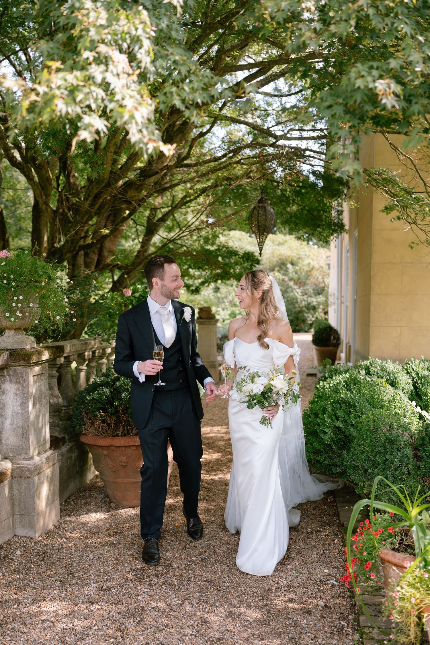 Newlyweds walking together under tree branches in a garden setting.