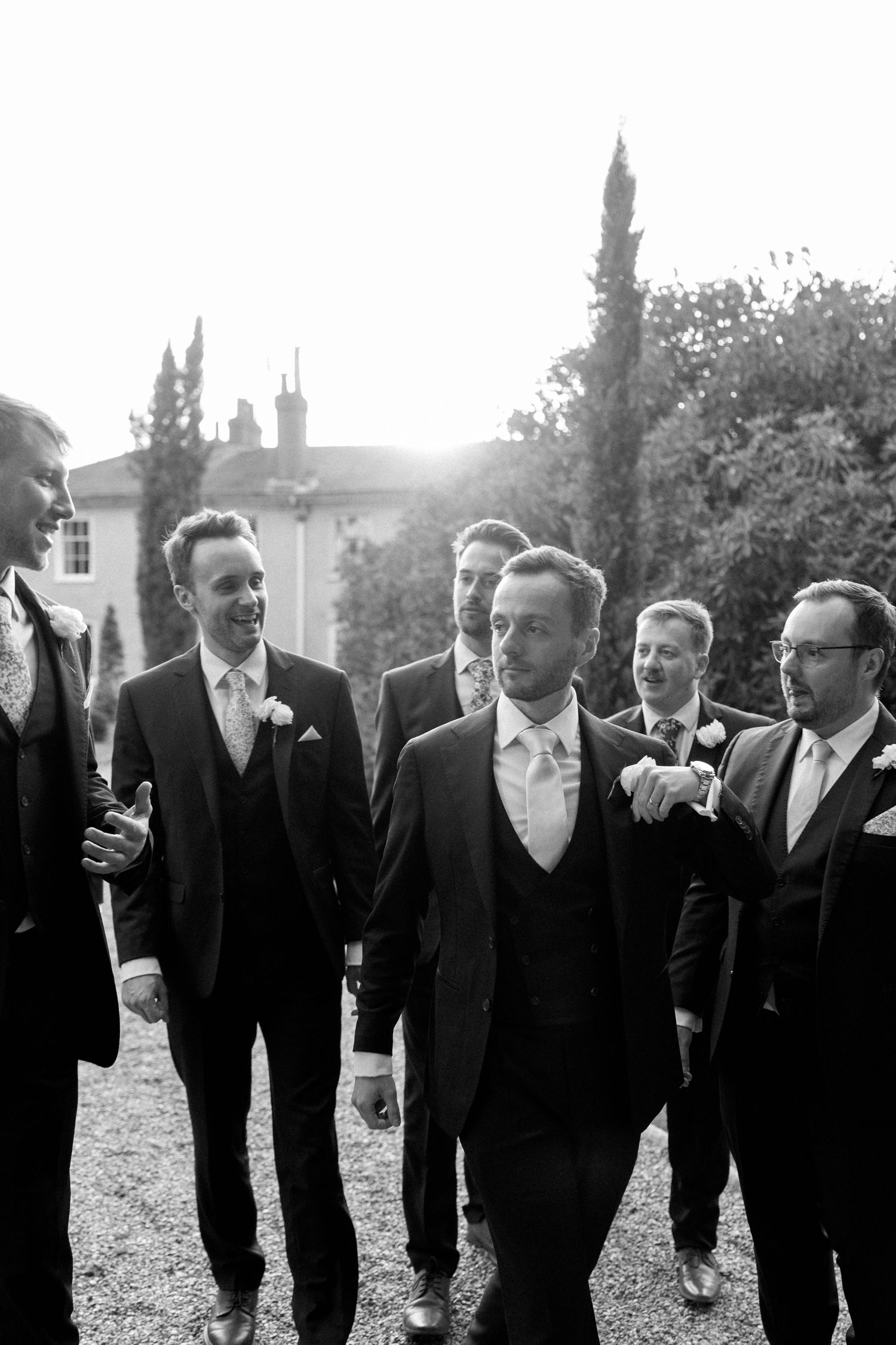 Groomsmen in formal black suits walking and laughing together on the grounds of a stately venue.