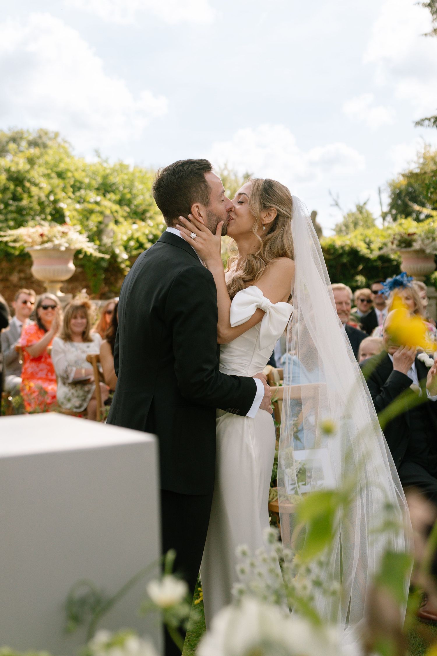 Romantic wedding kiss in a garden setting with guests and floral decorations.