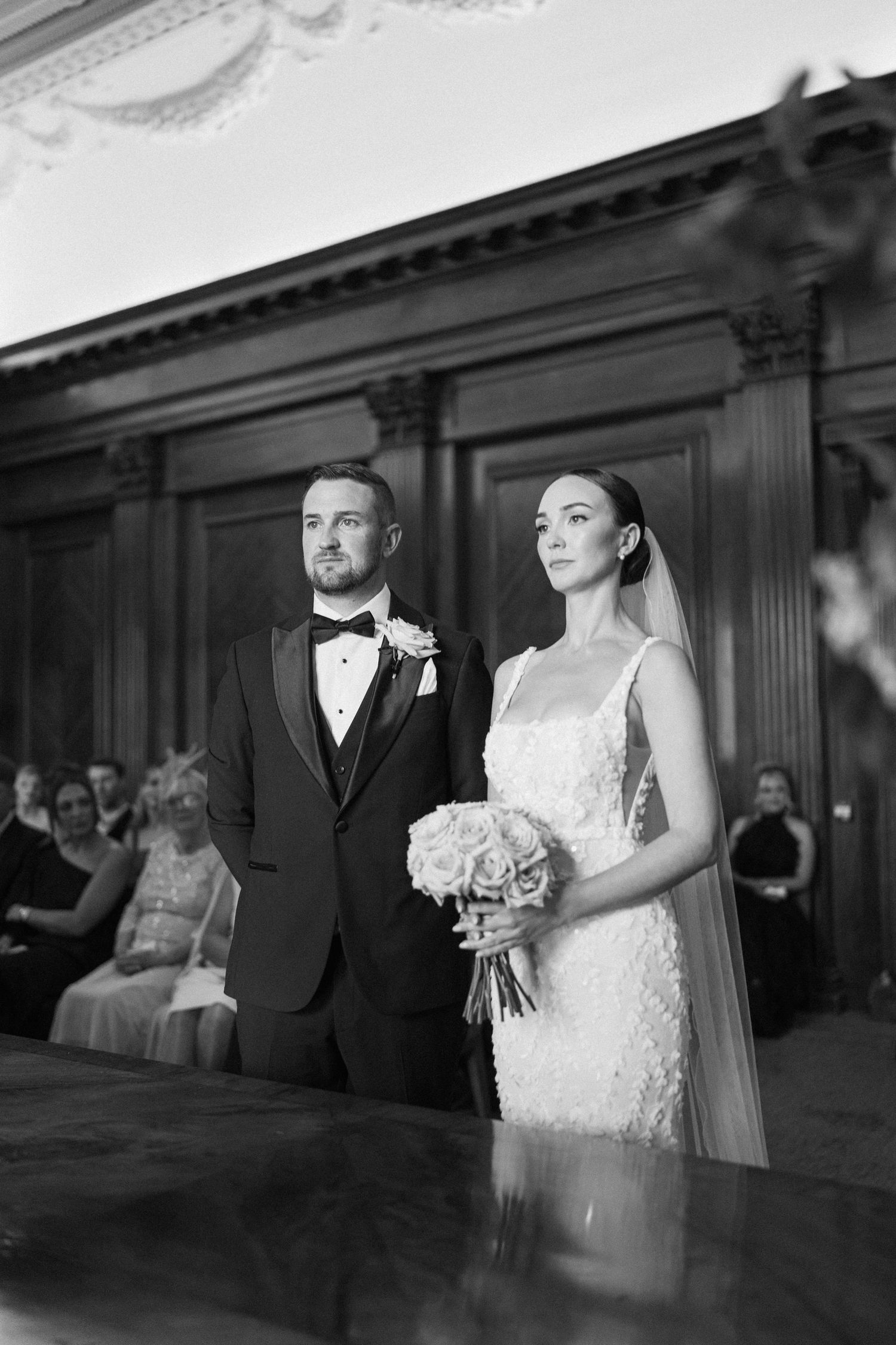A romantic black and white wedding photo showing the couple in formal attire against elegant wood paneling.