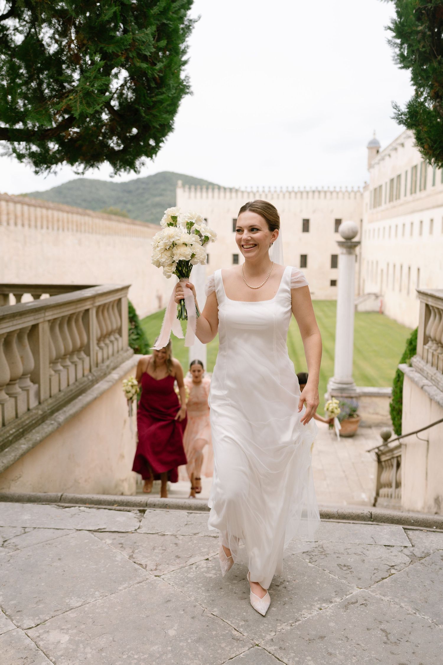 Wedding party walking up ornate stone staircase with castle and mountains in background.