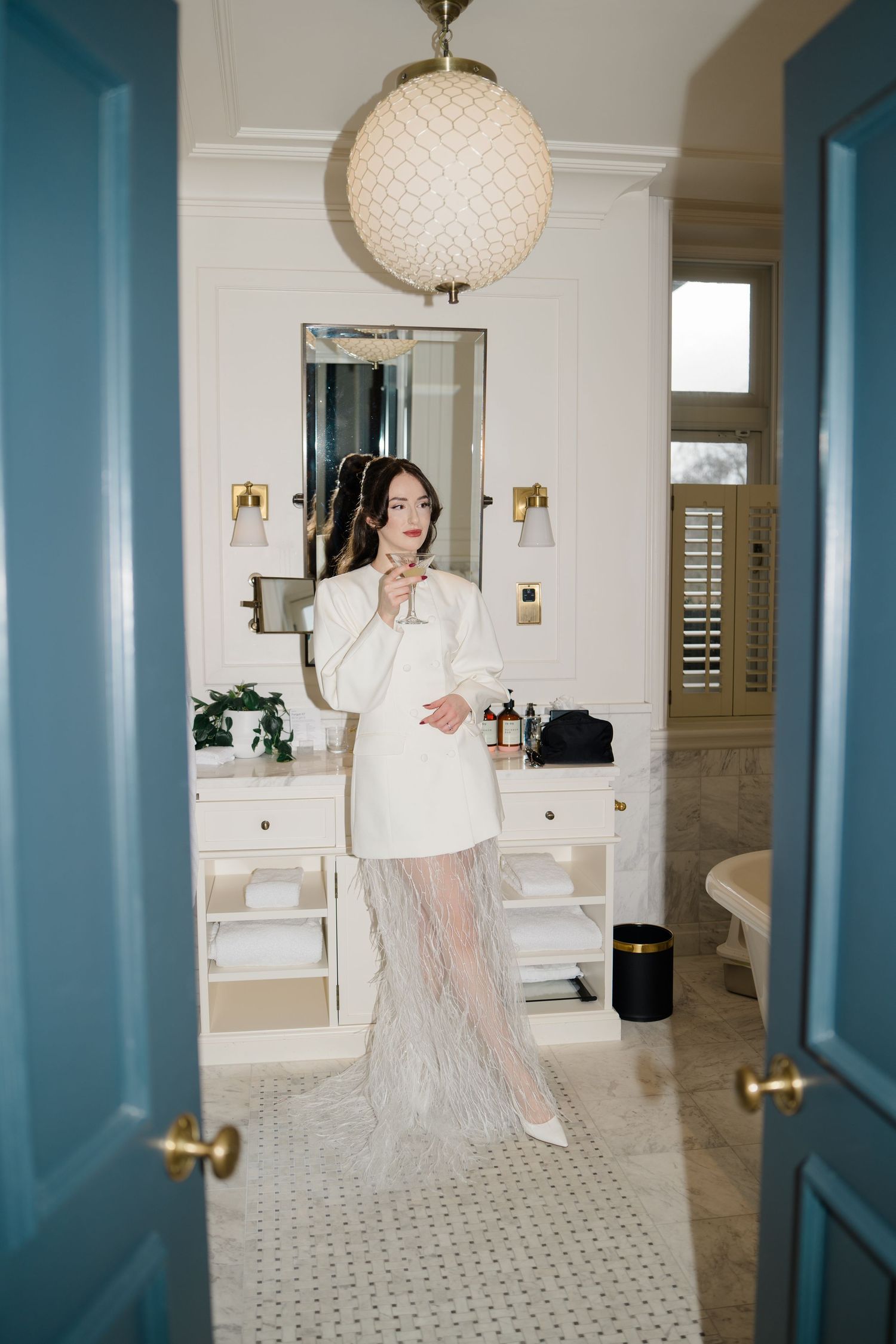 Bride in a white blazer and feather skirt posing in a luxurious white marble bathroom.
