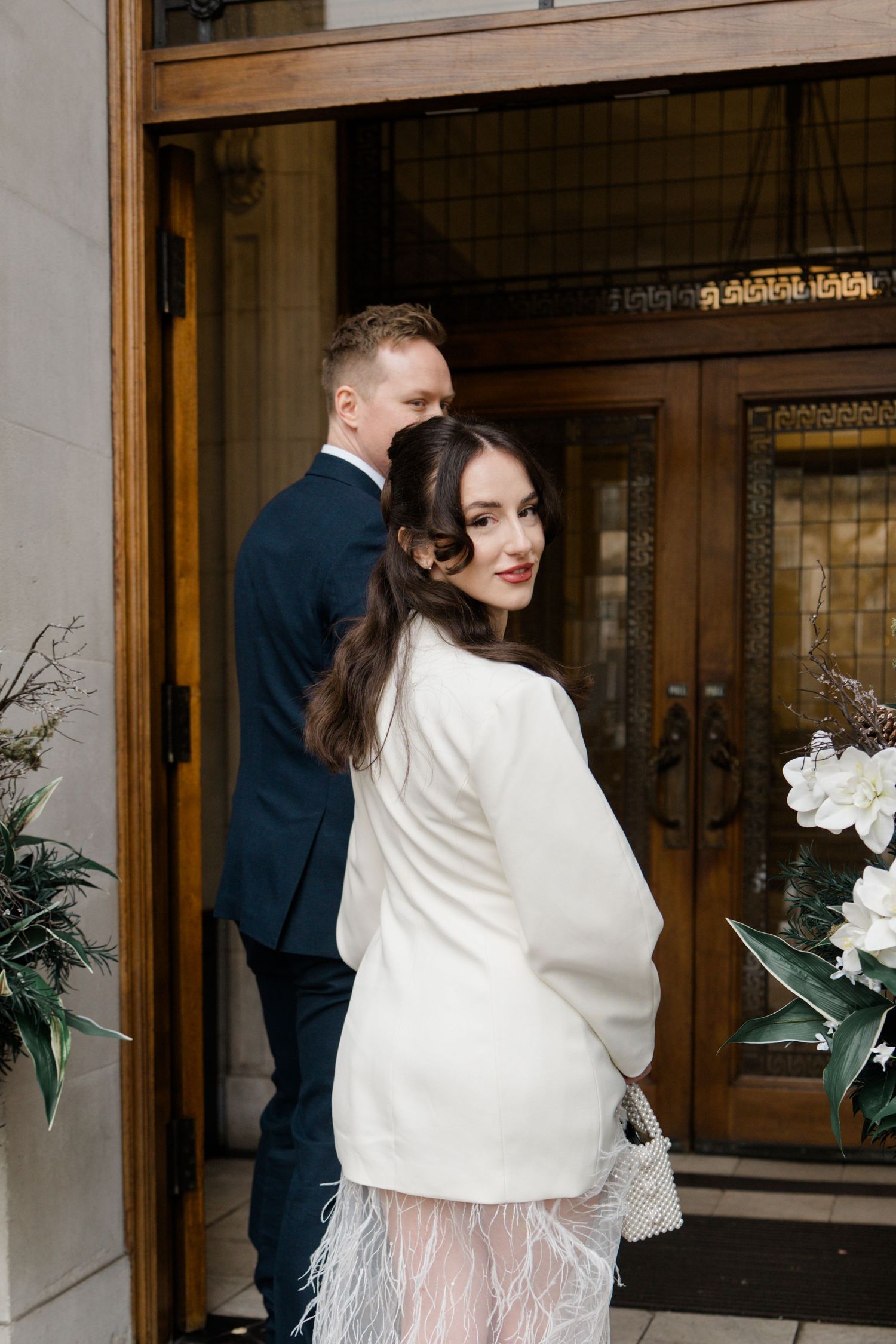 Smiling bride in white coat with feathered hem and groom entering ornate wooden doors.