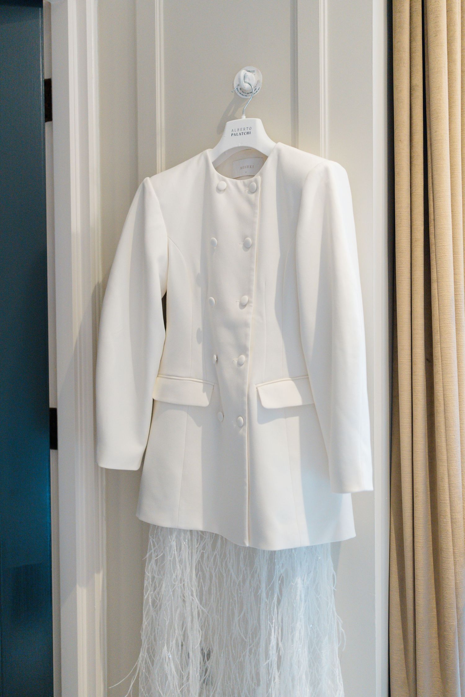White double-breasted bridal jacket with feather skirt hanging on door in elegant hotel room.
