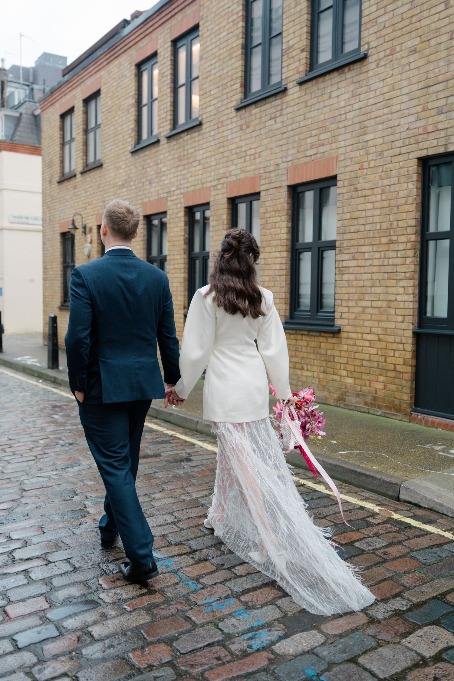 Bride in white blazer and feather skirt walking cobblestone street with groom.