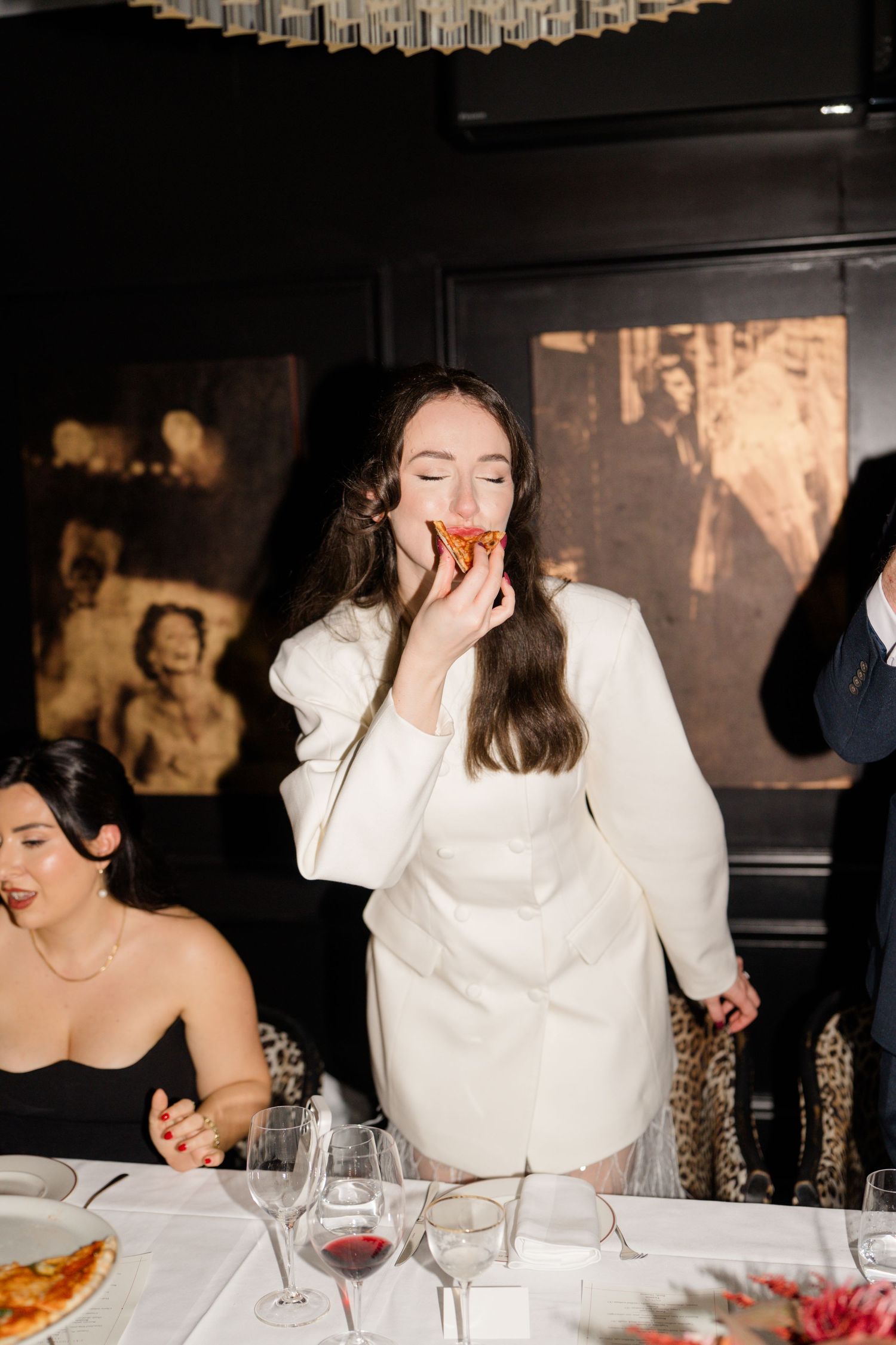 Bride in elegant white dress joyfully eating pizza at wedding reception dinner table with guests.