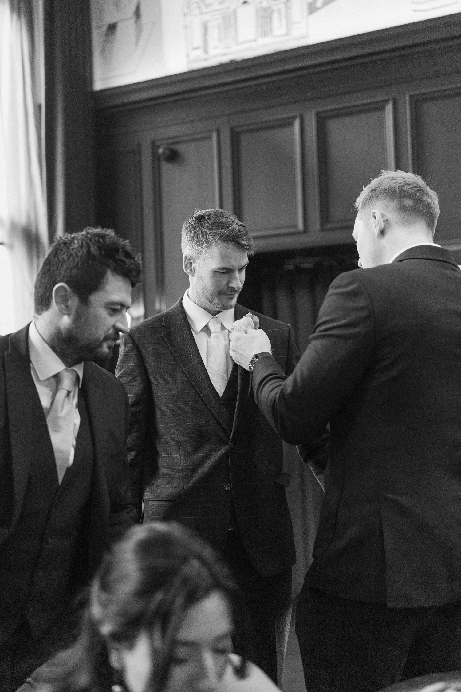 Black and white photo of groomsmen adjusting groom's boutonniere before wedding ceremony.