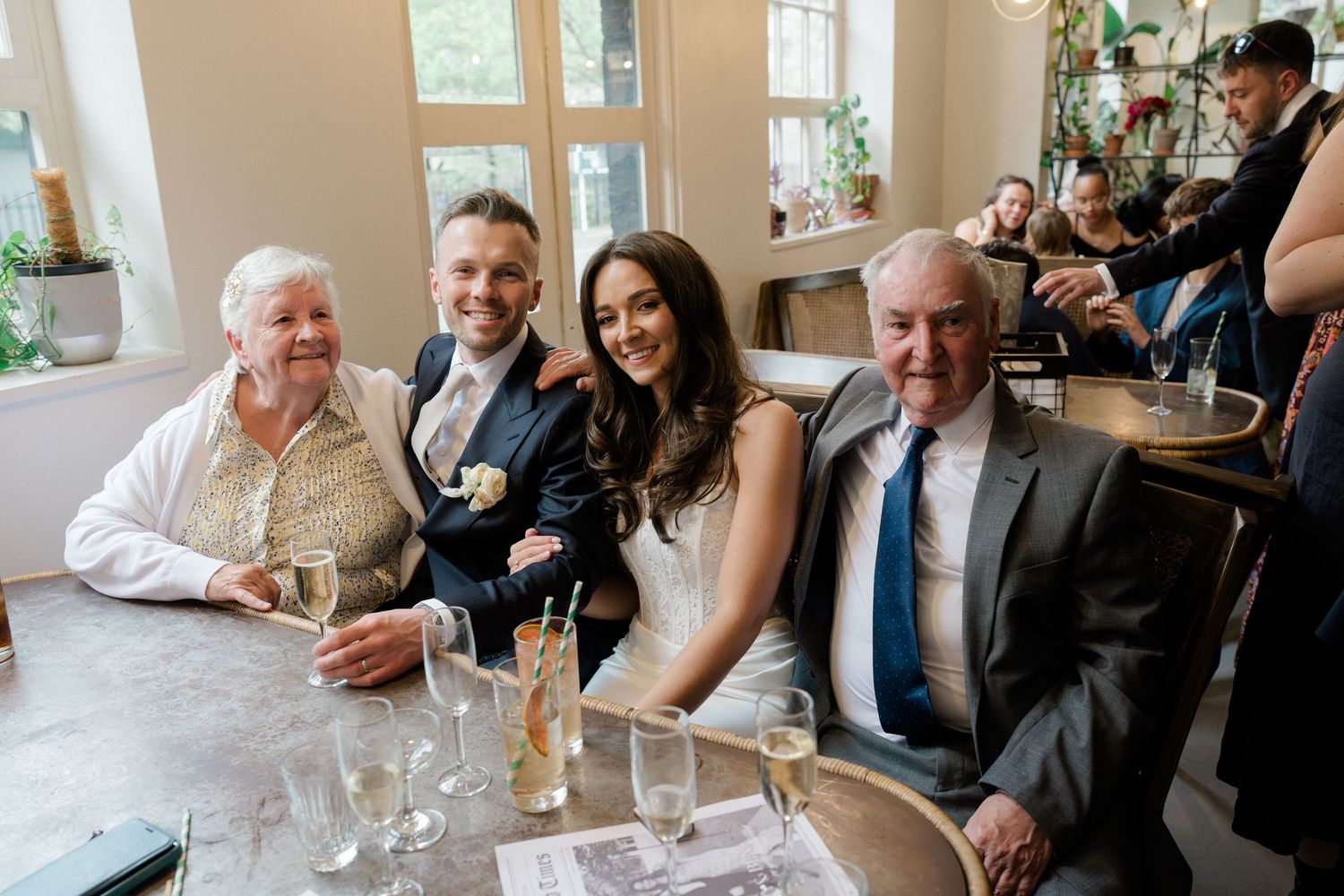 Bride and groom pose smiling with two older male guests at a wedding reception table with champagne glasses.