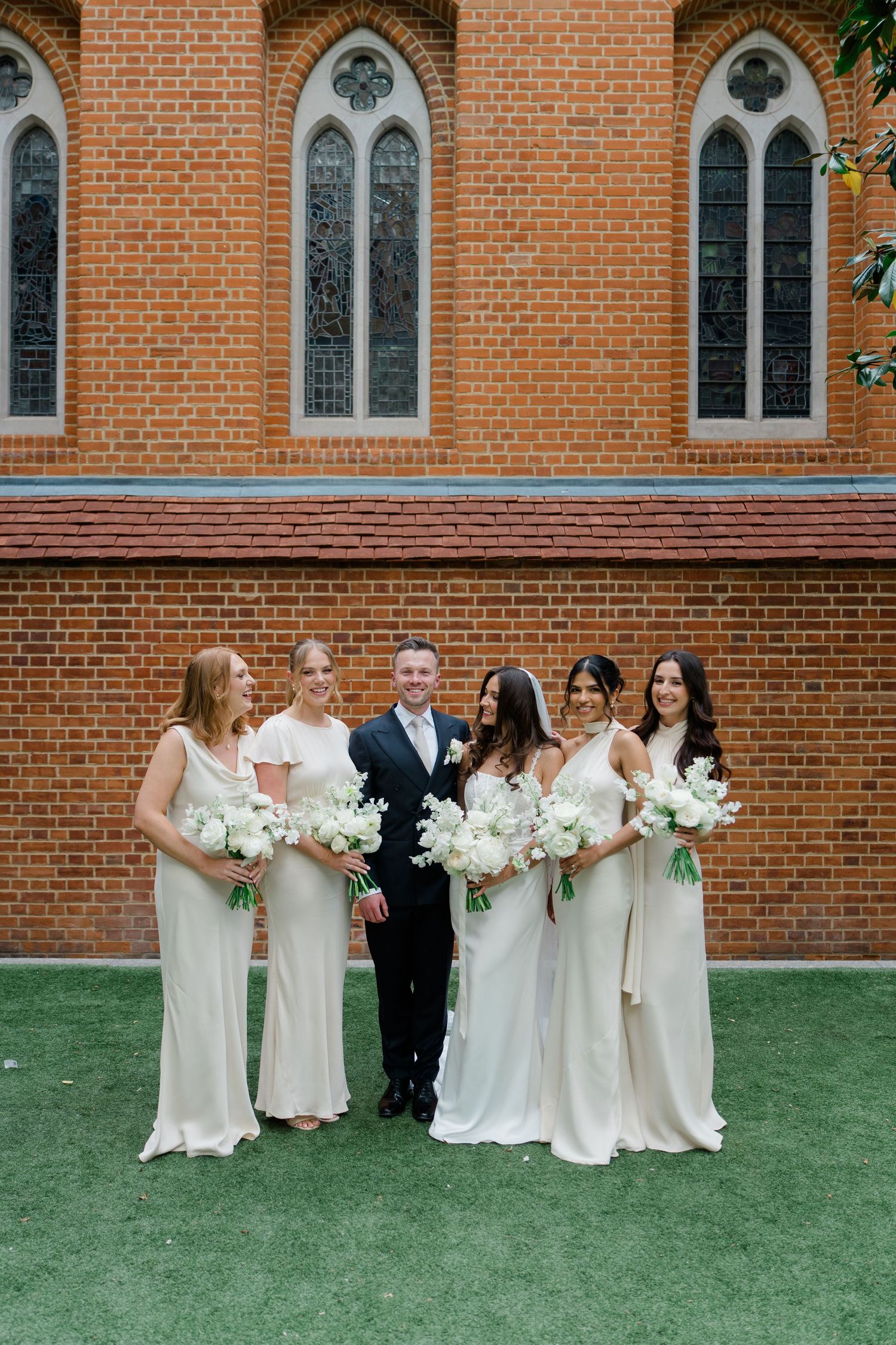 Groom in black suit poses with bride and four bridesmaids in cream gowns holding white bouquets.
