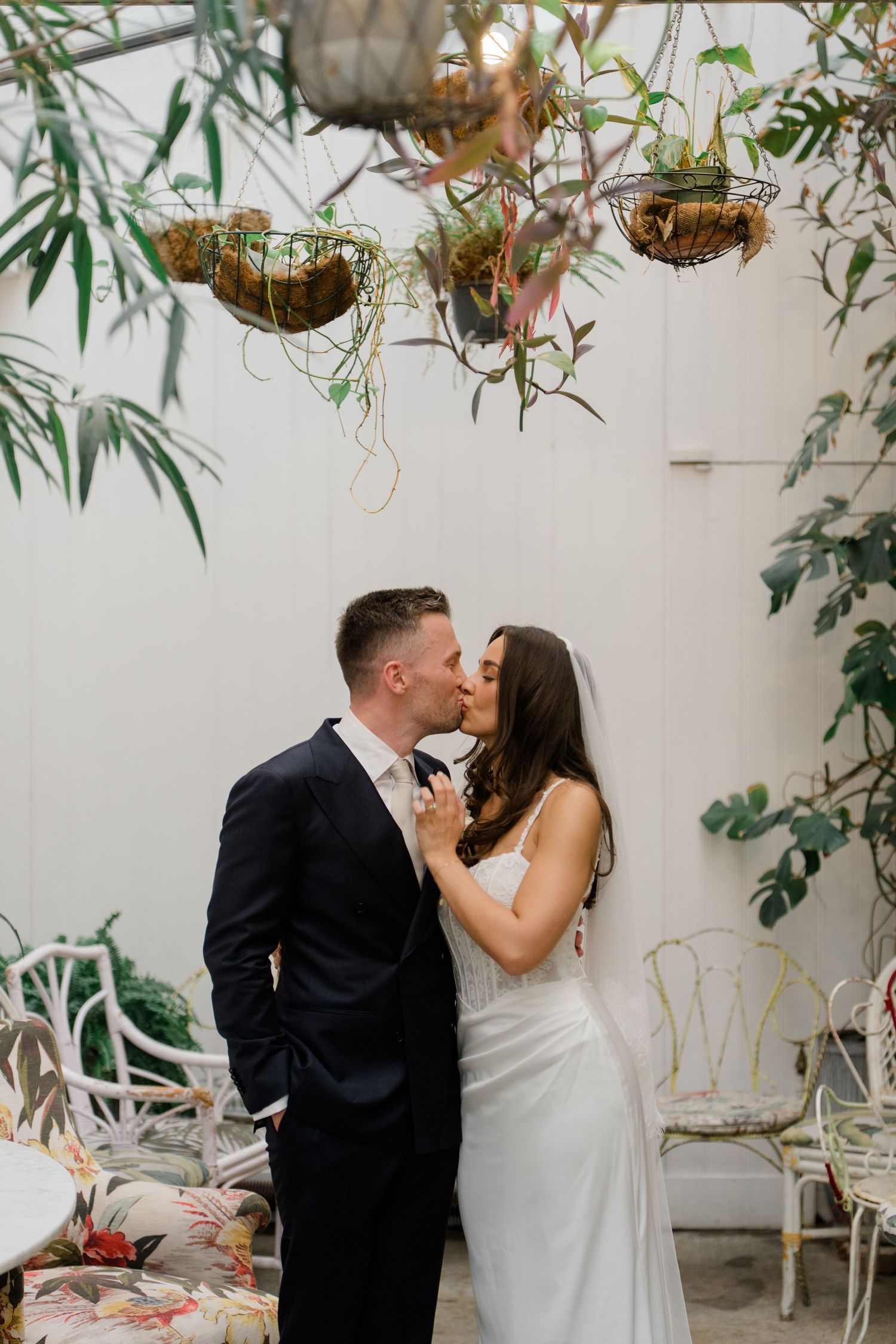 Bride and groom share a romantic kiss under hanging plants in a white botanical venue.