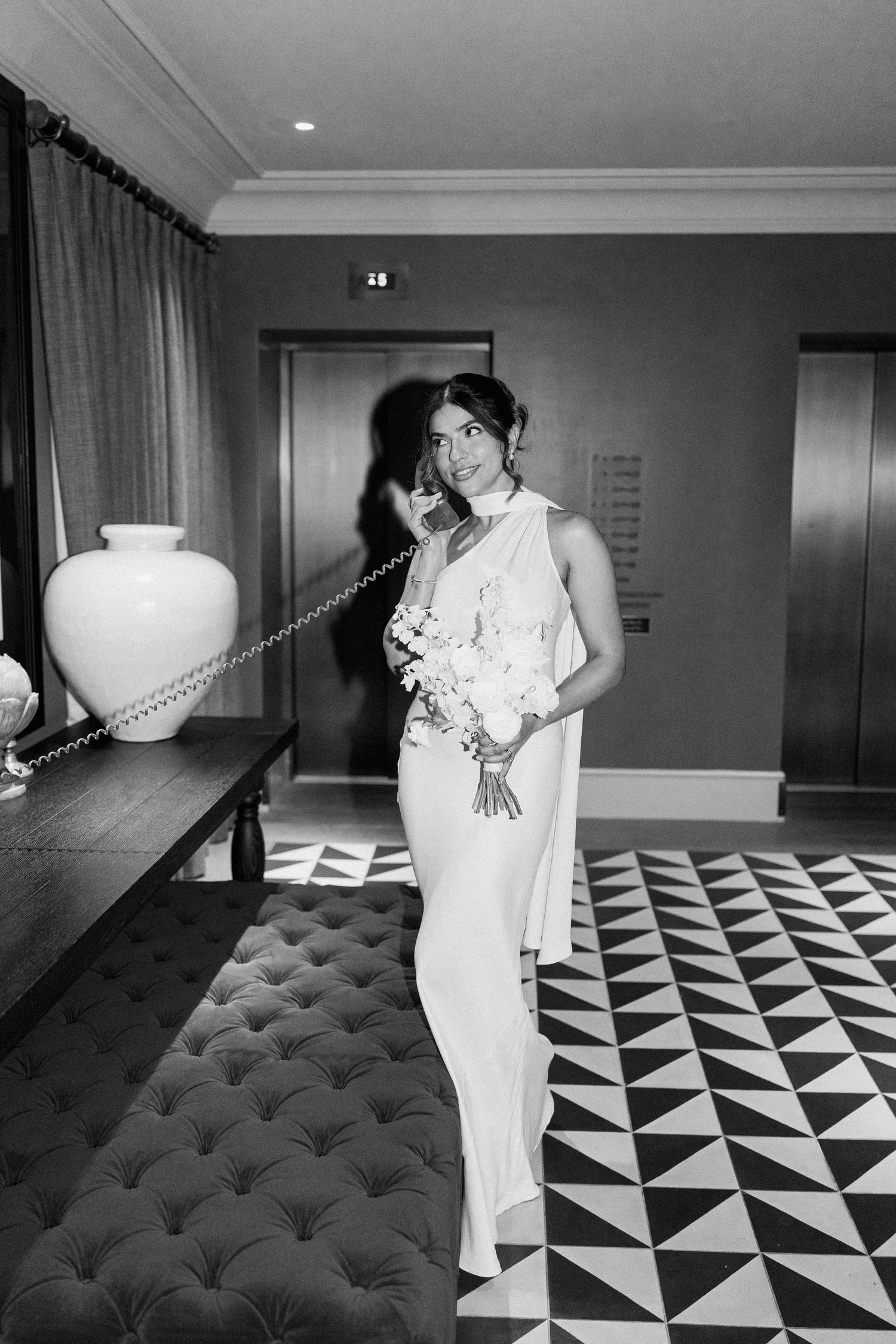 Elegant bride in a sleek white one-shoulder gown holding a white bouquet in a luxury hotel lobby, black and white photo.