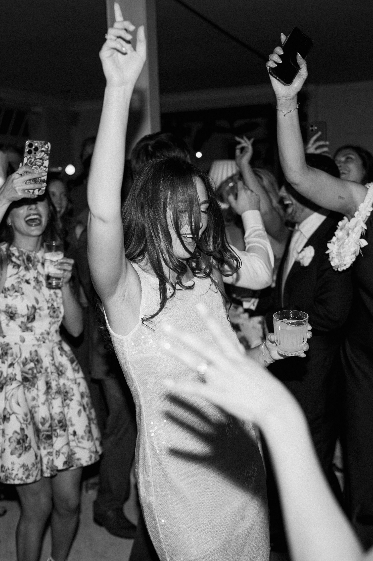 Black and white photo of bride in sequin dress dancing with arms raised at lively wedding reception.
