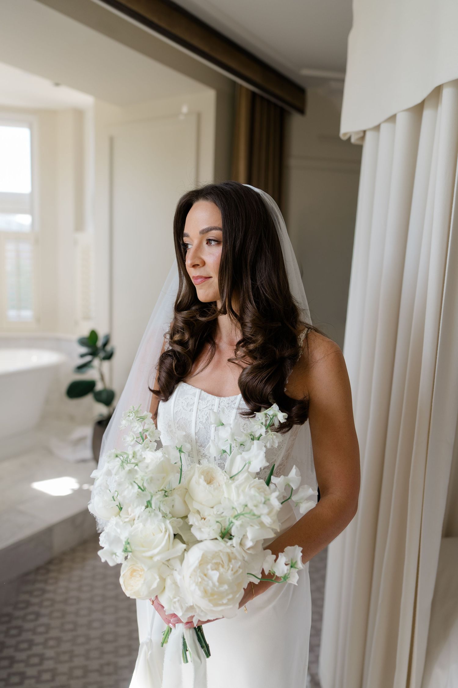 Elegant bride in white lace gown and veil holding white peony bouquet by a bright window.