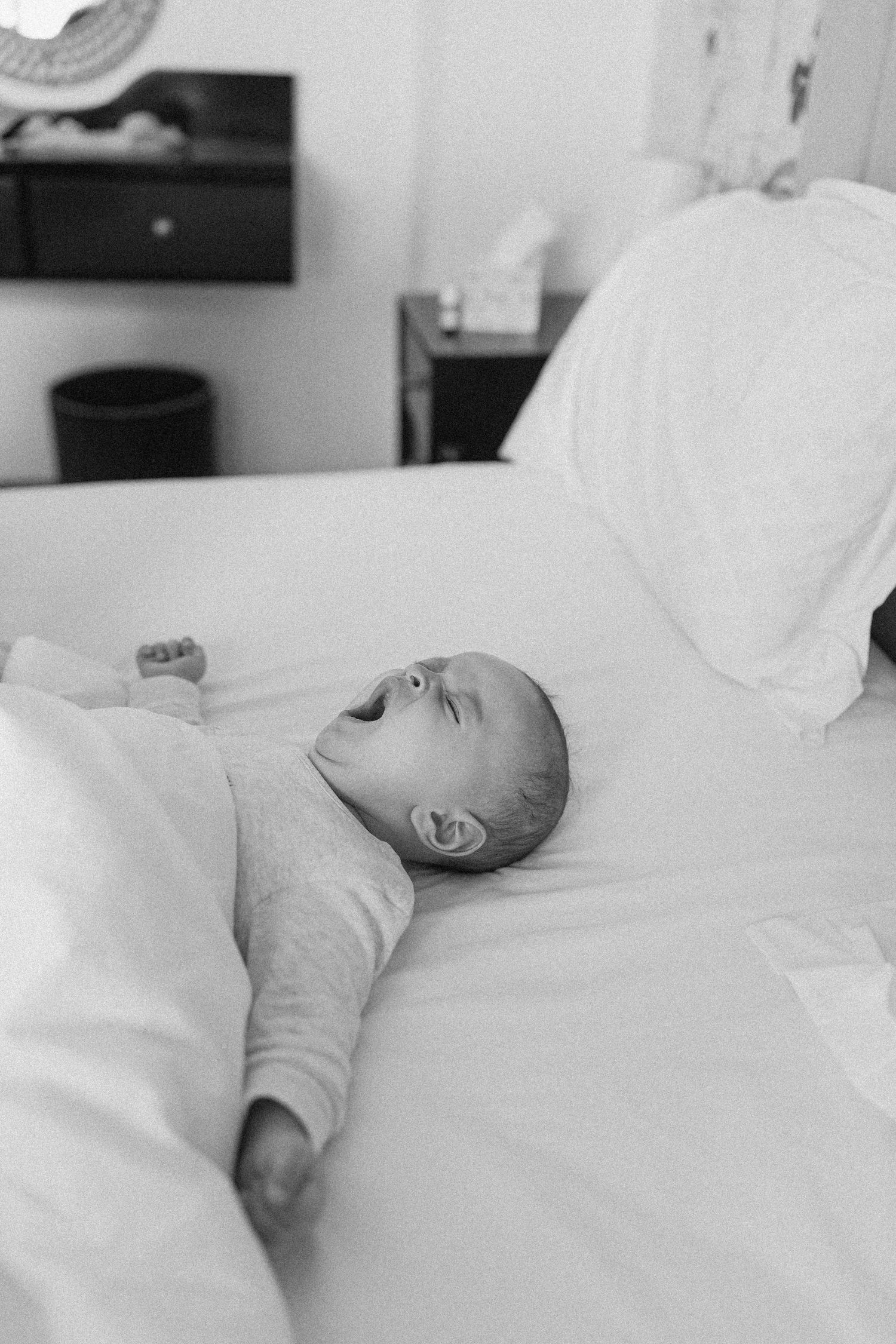 Black and white photo of a yawning newborn baby lying on a white bed, captured in a cozy bedroom setting.
