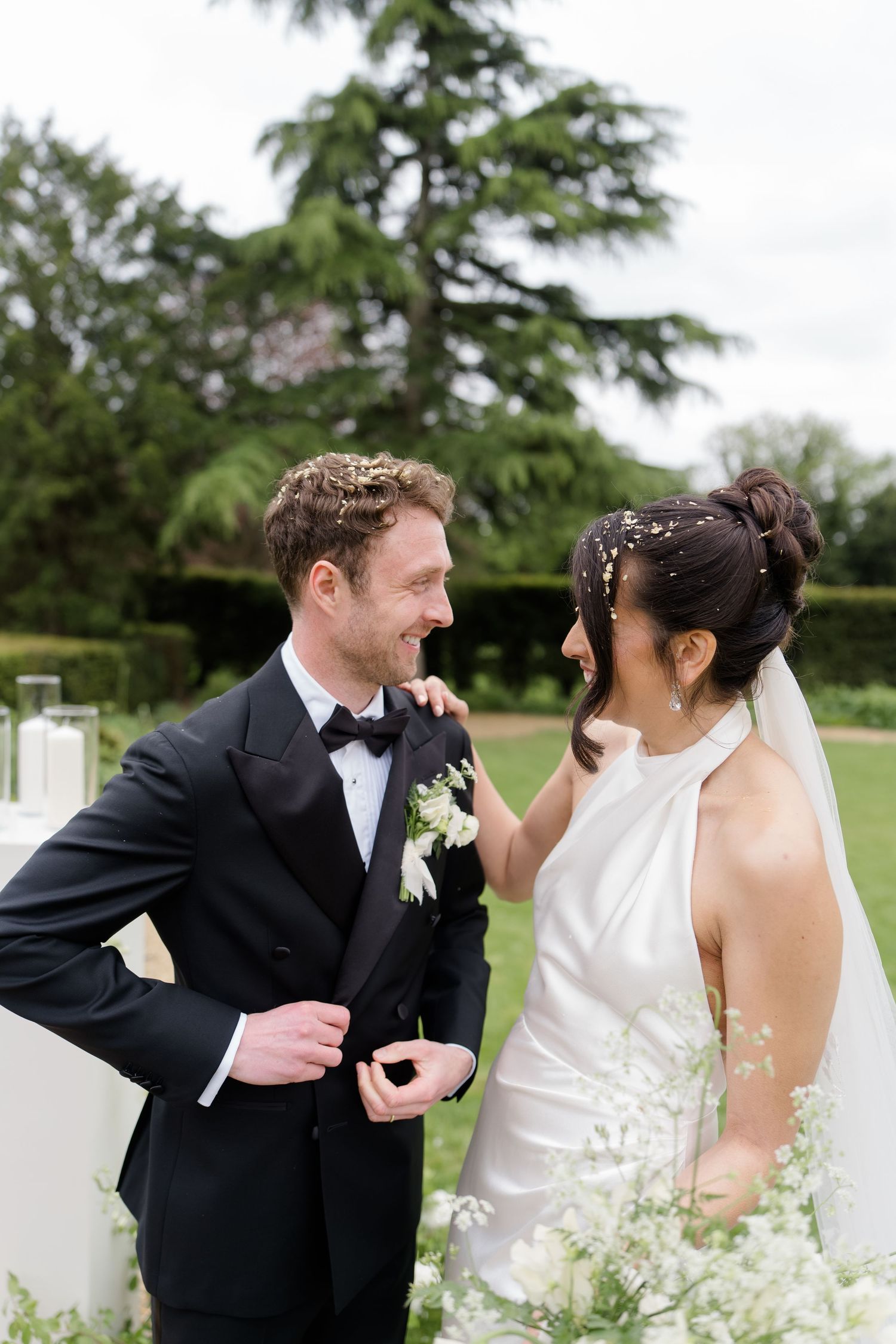Bride and groom share a joyful moment during outdoor wedding ceremony, bride in halter gown touching grooms tuxedo lapel.