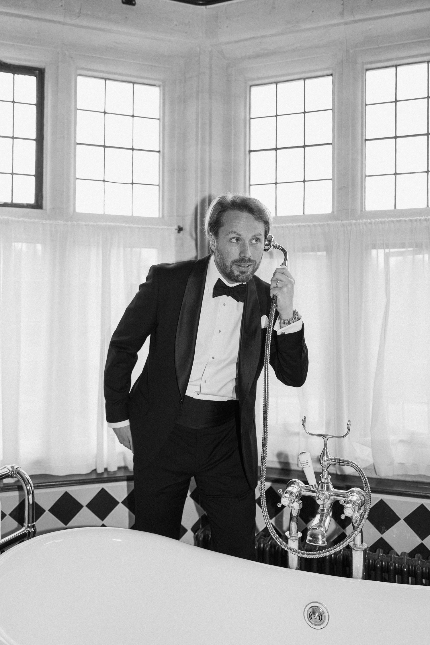 Black and white photo of groom in tuxedo holding shower head beside a freestanding bathtub in bay window bathroom.