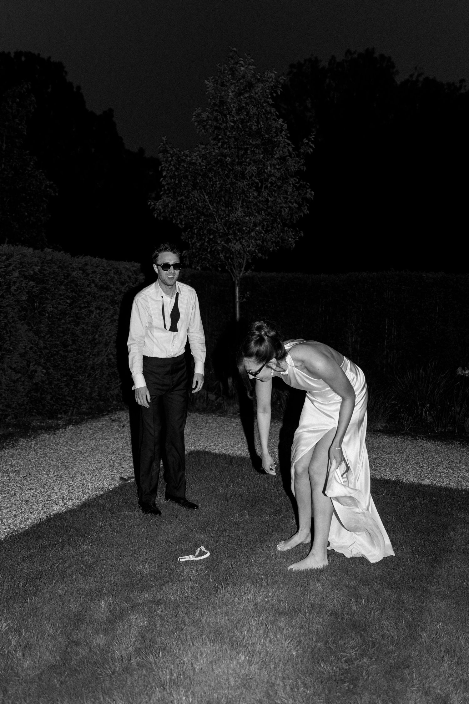 Black and white photo of groom watching barefoot bride bend down in garden at night.