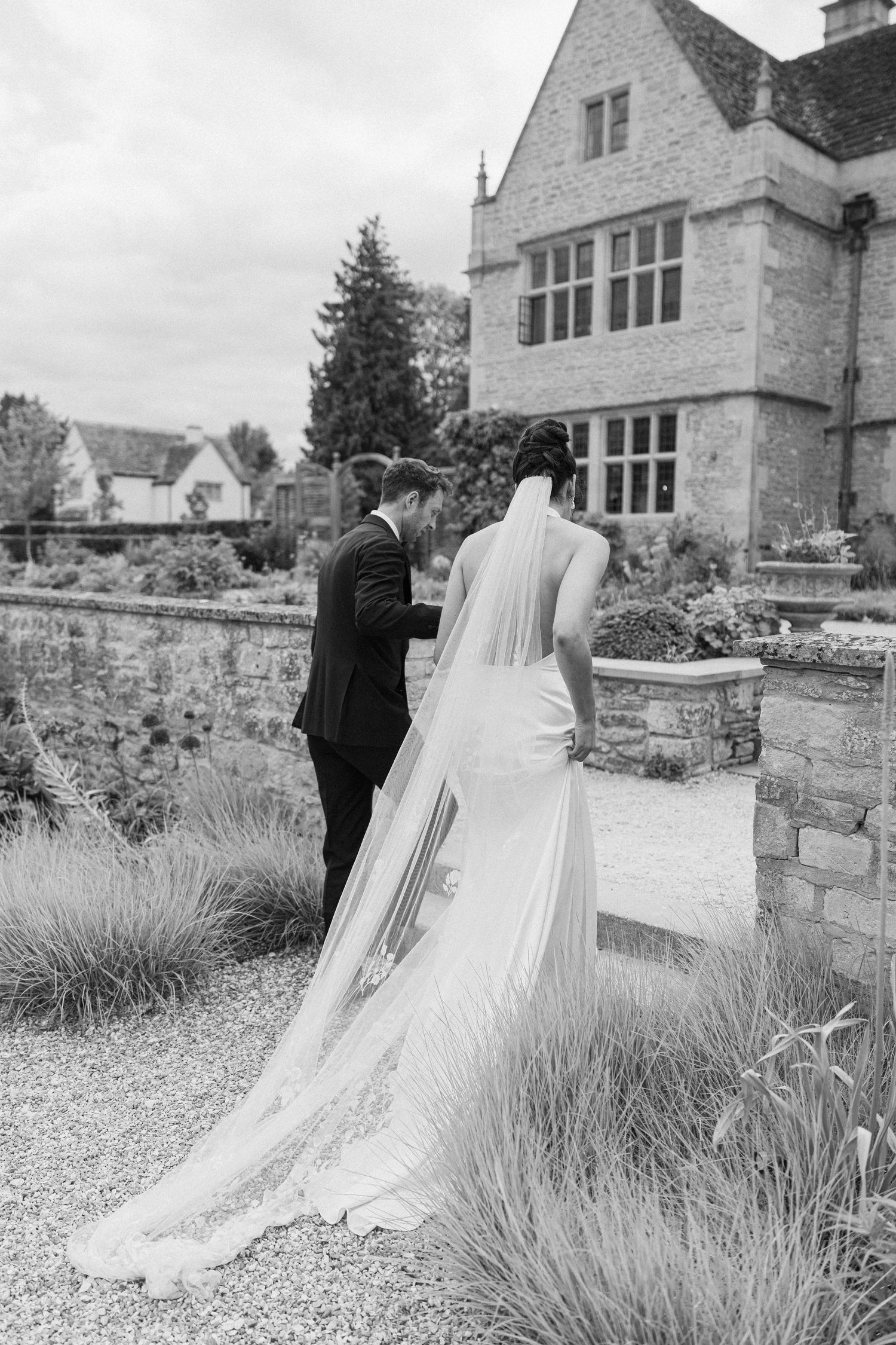 Black and white photo of bride and groom walking through elegant manor garden, brides cathedral veil trailing on gravel path.
