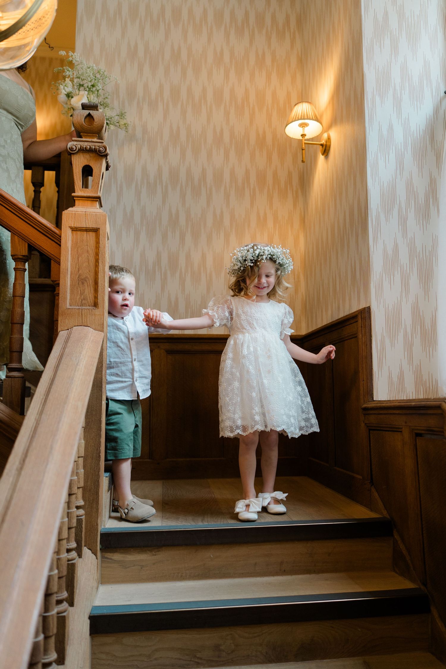 A flower girl in a white lace dress and floral crown walks down wooden stairs with a young ring bearer at a wedding.