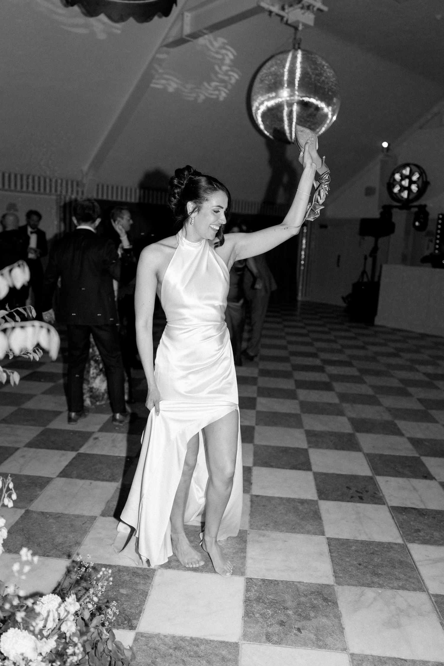 Black and white photo of bride in halter gown dancing under disco ball on checkered dance floor.