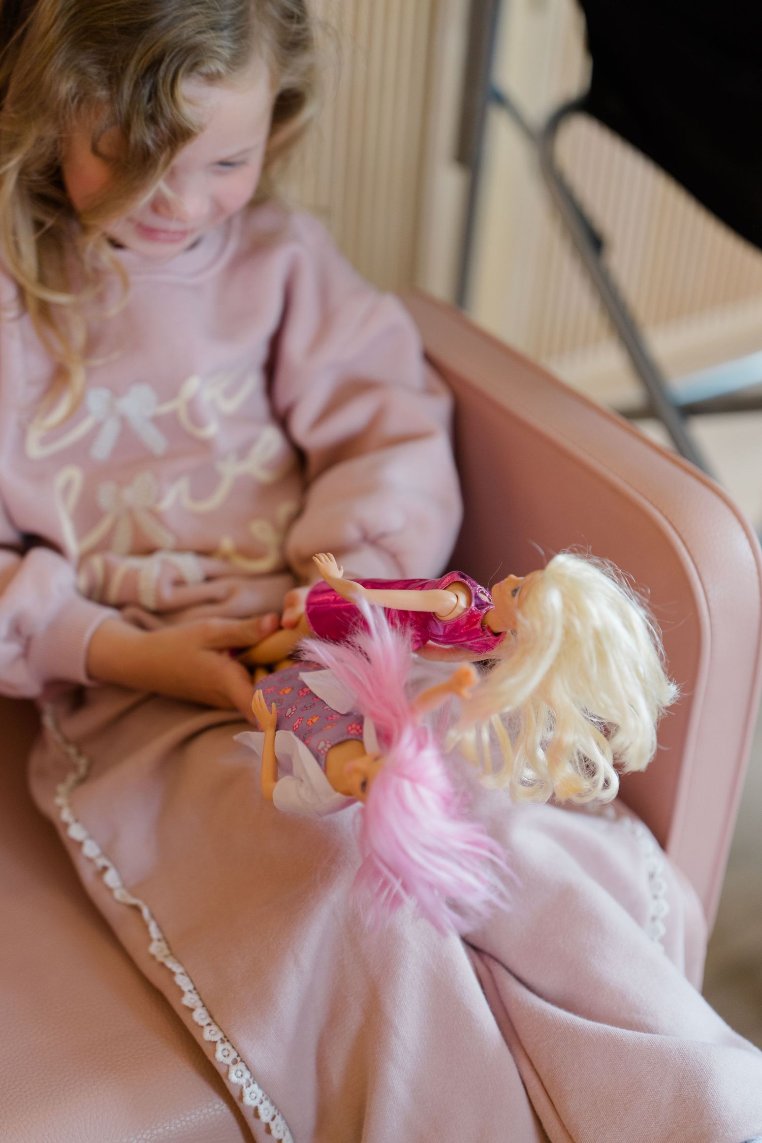 Young girl in pink sweatshirt playing with Barbie dolls while seated in a pink chair.