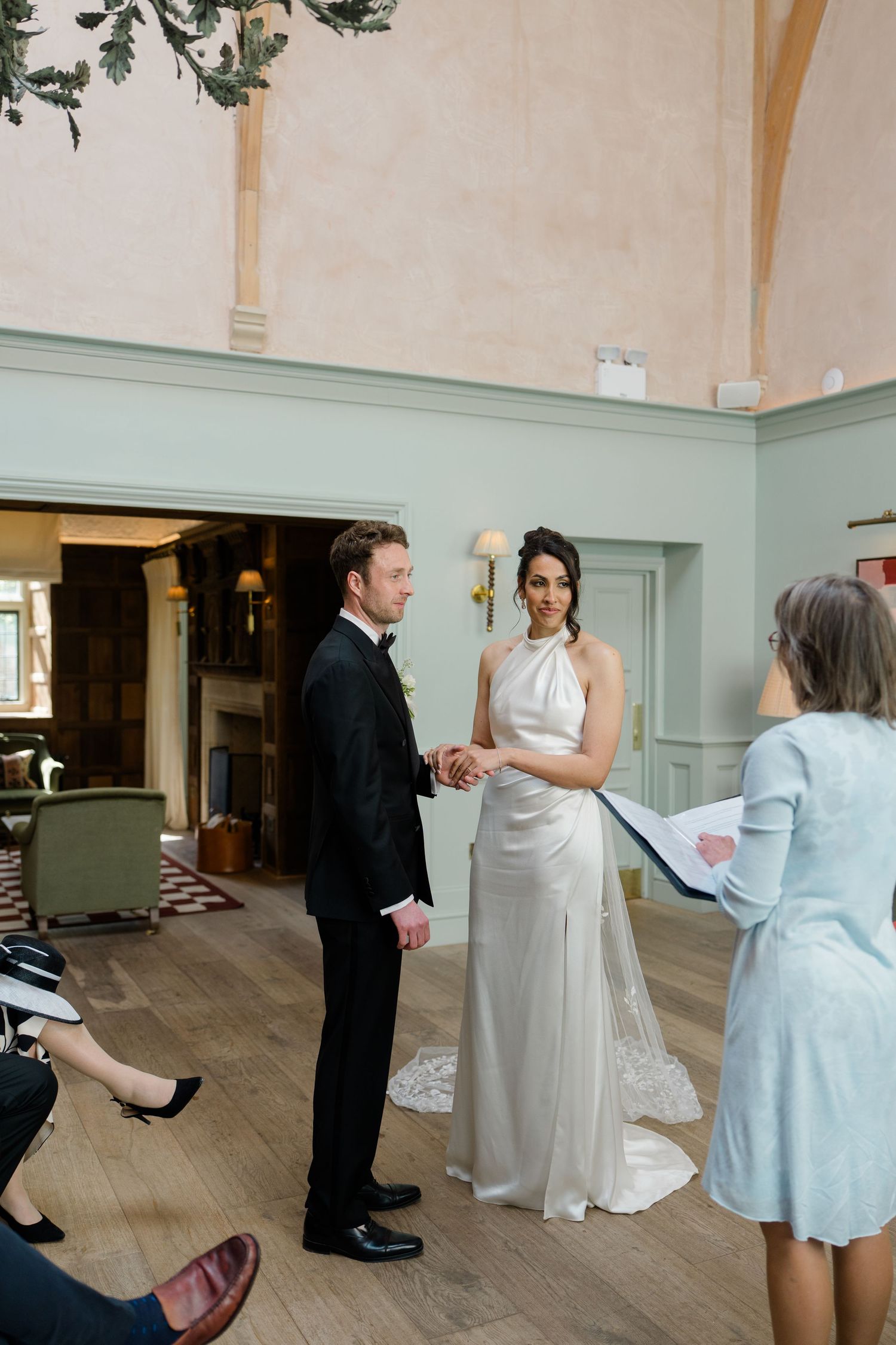 Bride and groom hold hands during wedding ceremony vows in an elegant manor house with sage green paneled walls.