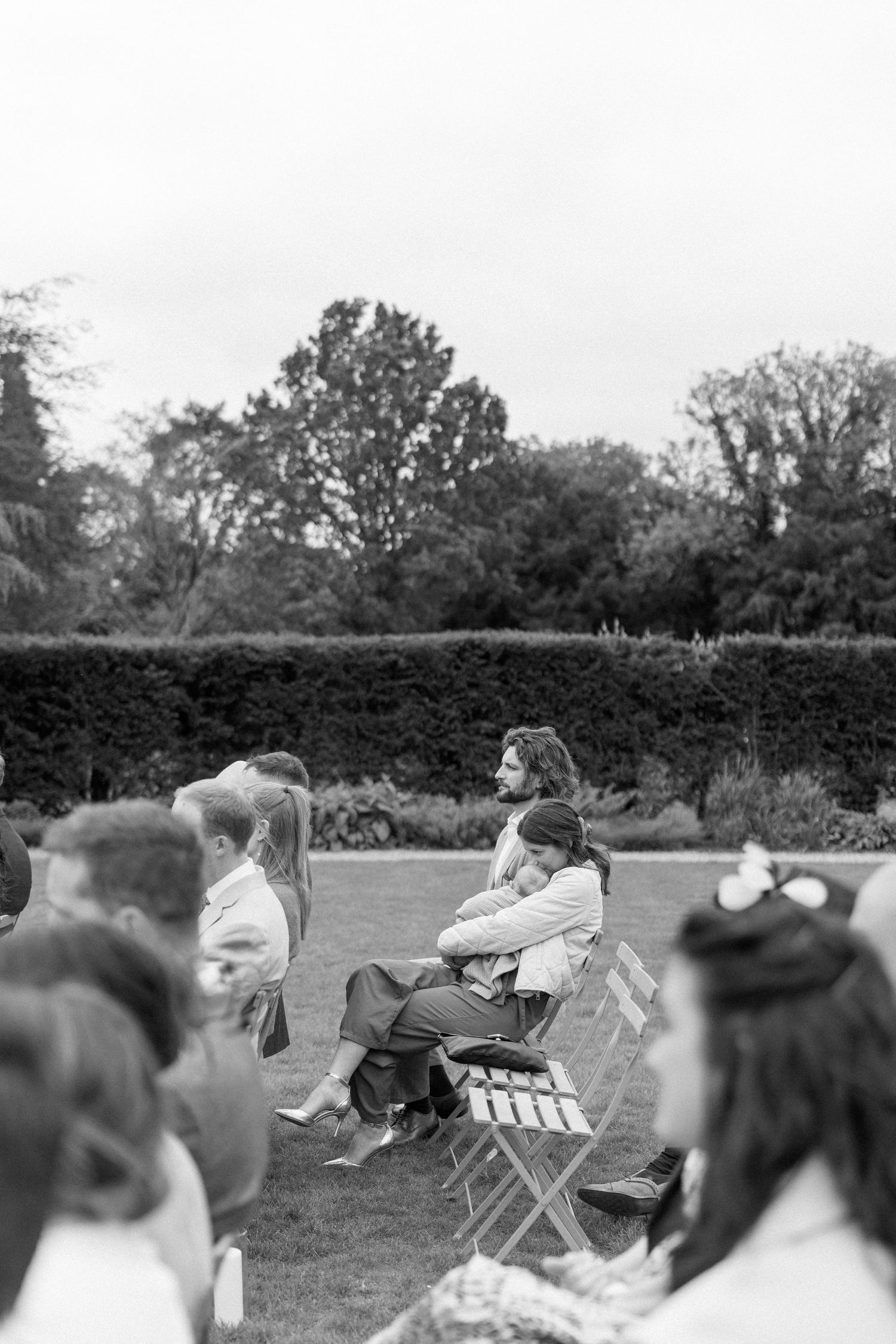 Guests seated on wooden chairs outdoors during a garden wedding ceremony, surrounded by lush hedges and trees.