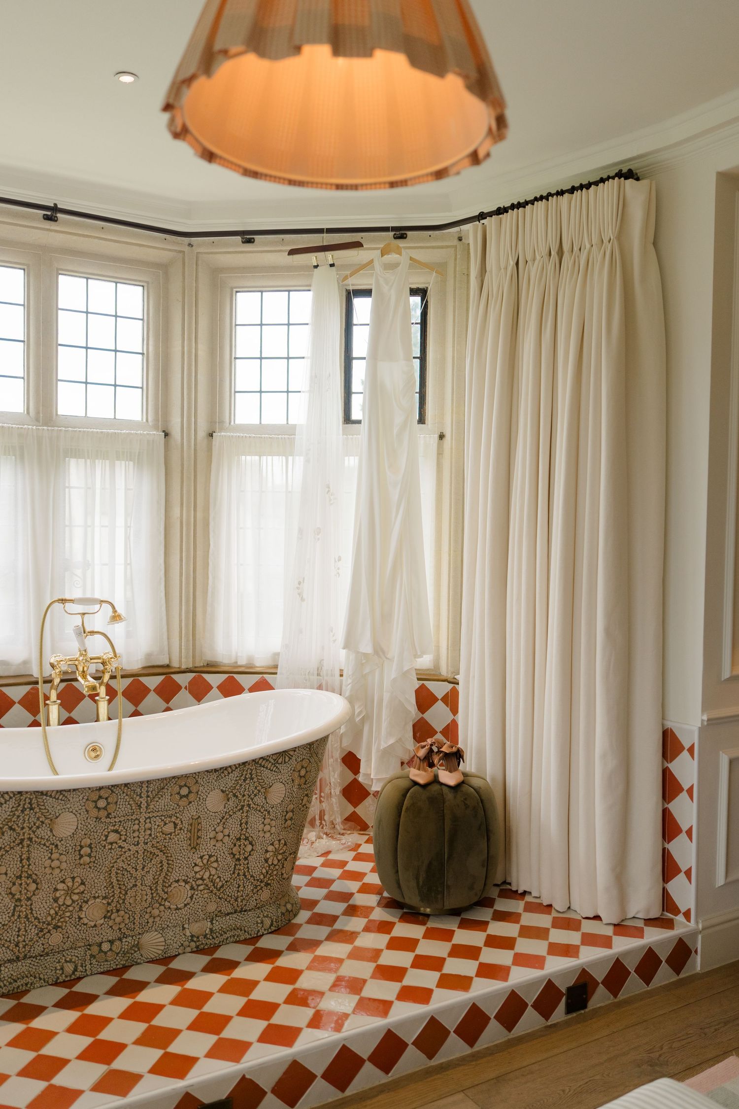 Elegant bathroom with ornate freestanding bathtub, red checkered floor tiles, and wedding dress hanging by window.
