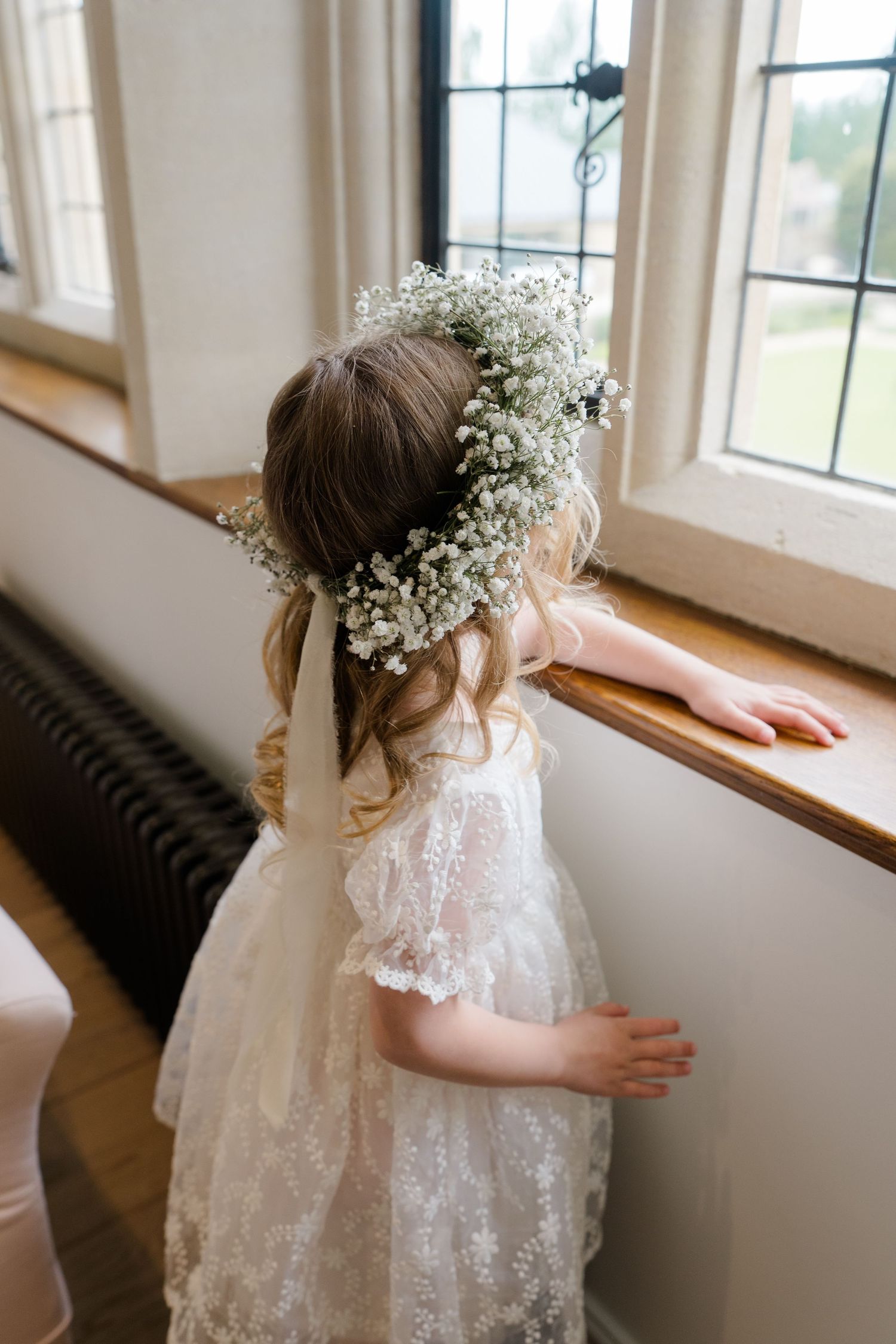 A flower girl in lace dress wearing a baby's breath floral crown gazes out a window.