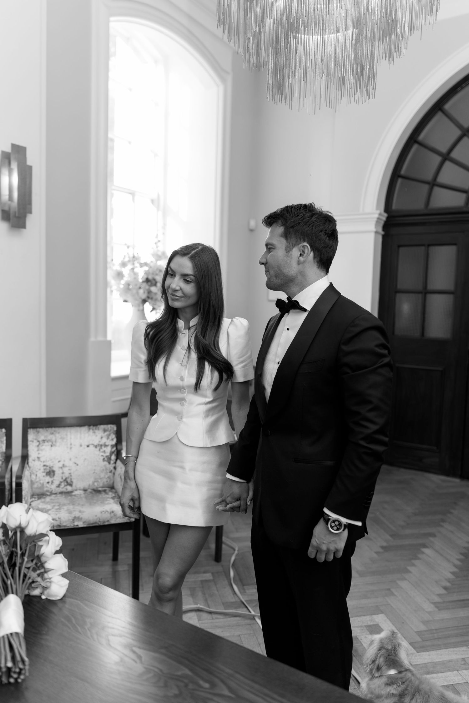 Black and white photo of bride in white mini dress and groom in tuxedo holding hands indoors.