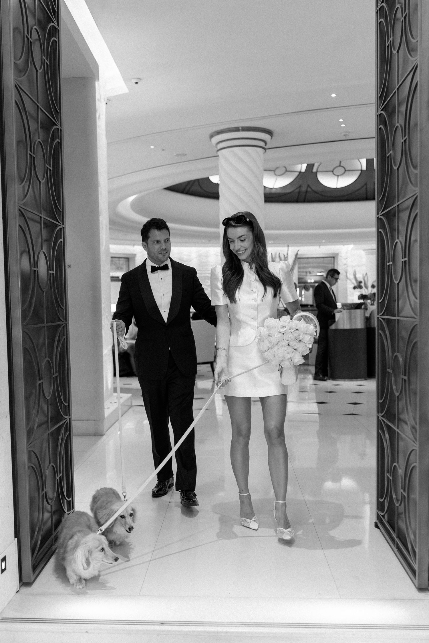 Bride and groom walk with a corgi dog through an elegant hotel lobby in black and white photo.