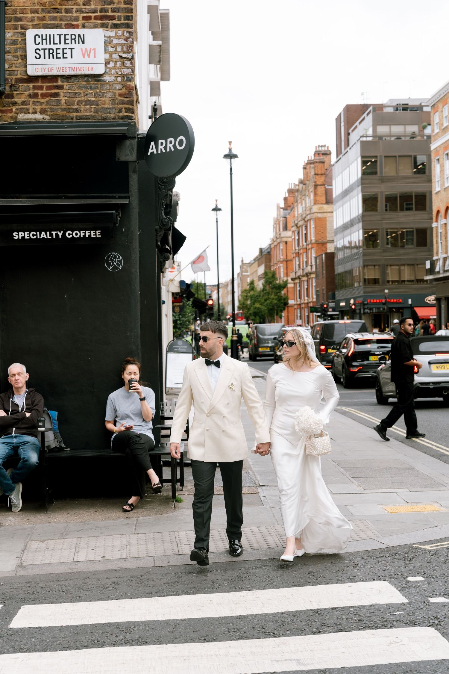 A couple in white attire crosses Whitfield Street near an Arro coffee shop in London.