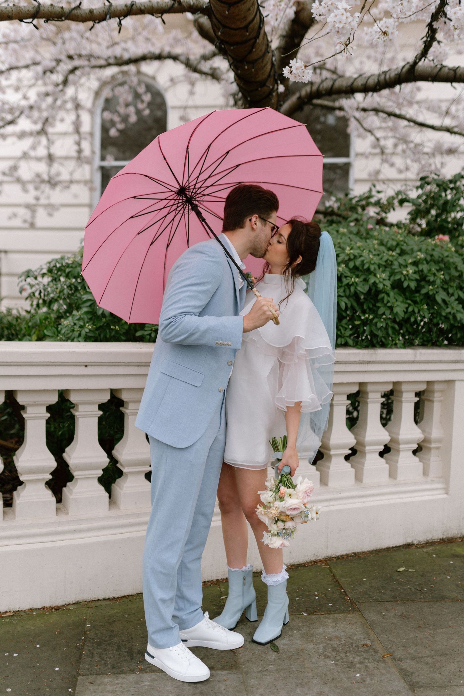 A couple shares a romantic moment under a pink umbrella near white stone railings with cherry blossoms overhead.