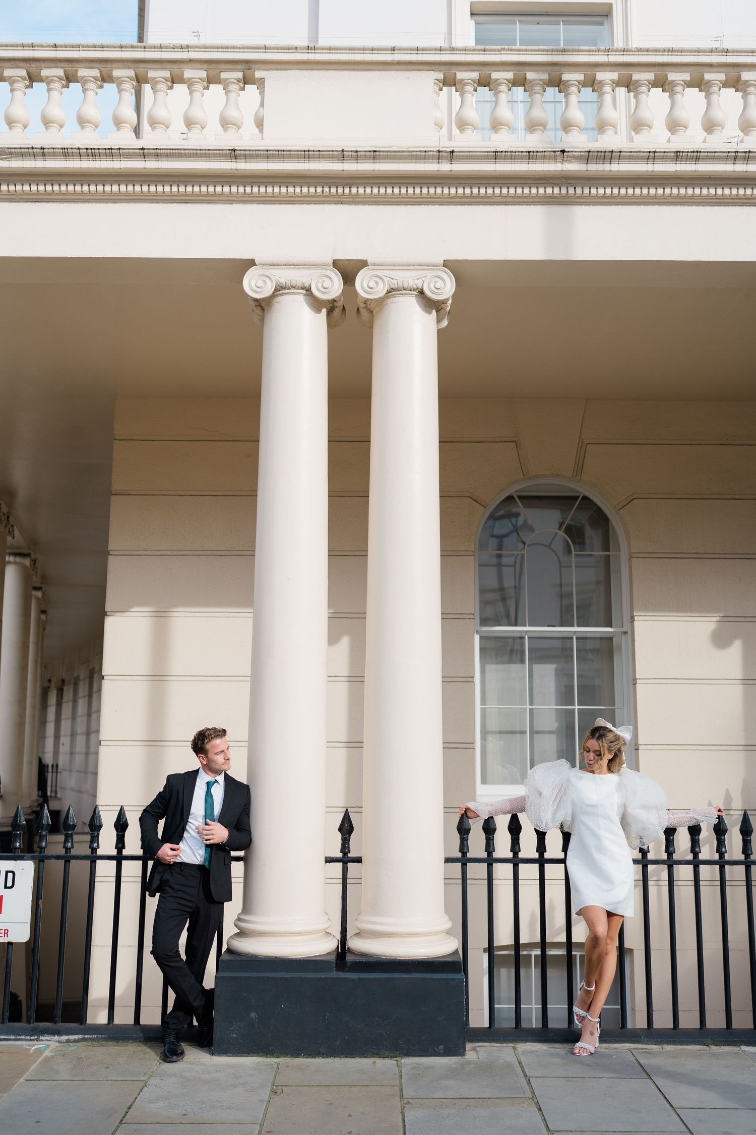 Playful couple interacts between classical white columns and iron fencing.