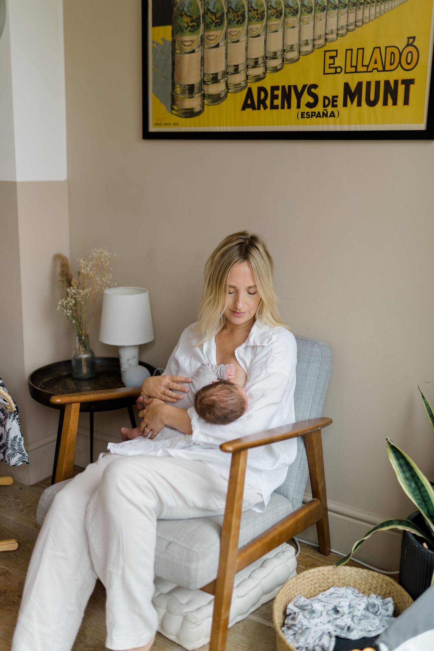 A peaceful nursery scene with a mid-century armchair, vintage poster, and intimate feeding moment in white decor.