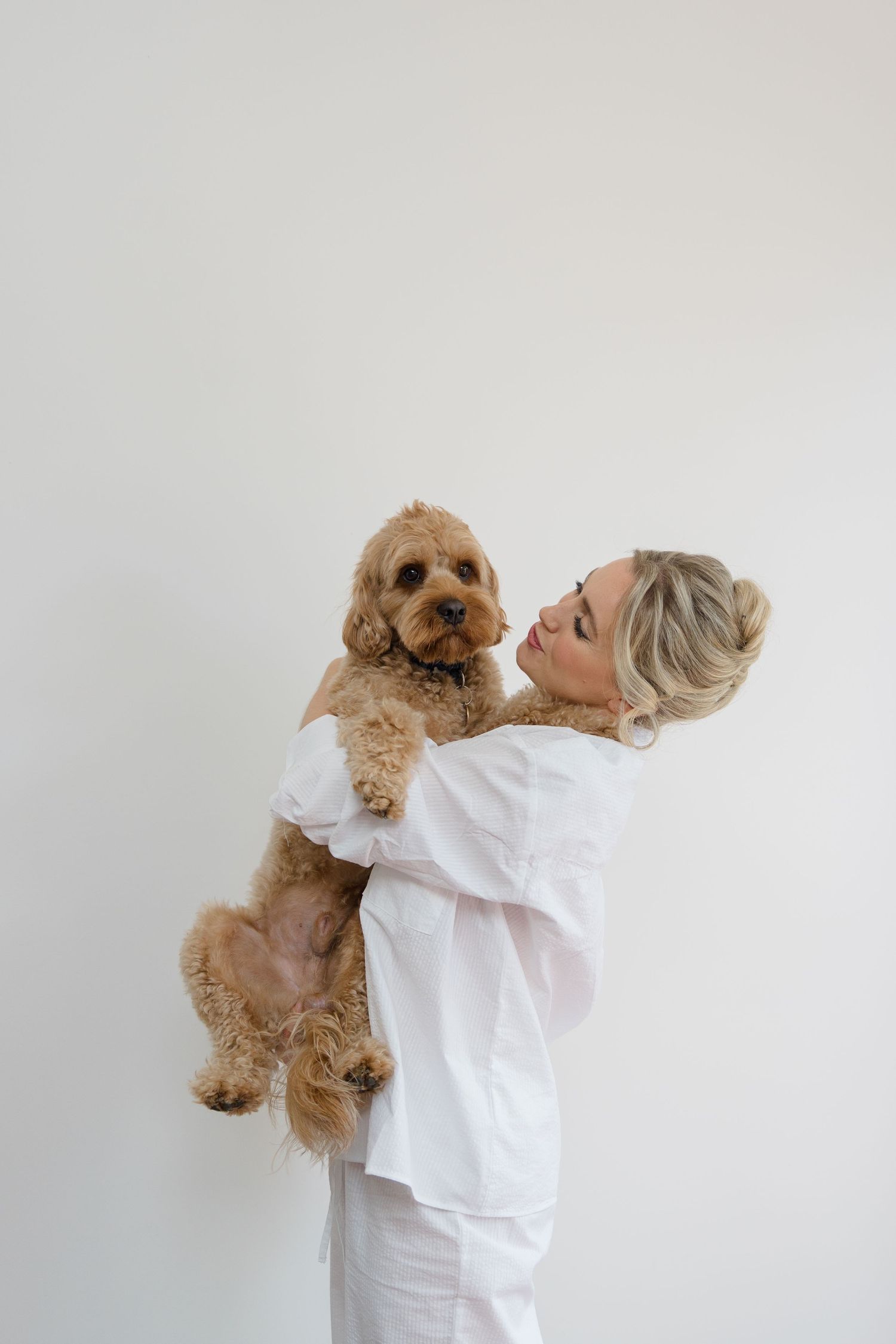 A person in white clothing holds and cuddles an adorable golden doodle dog against a plain background.