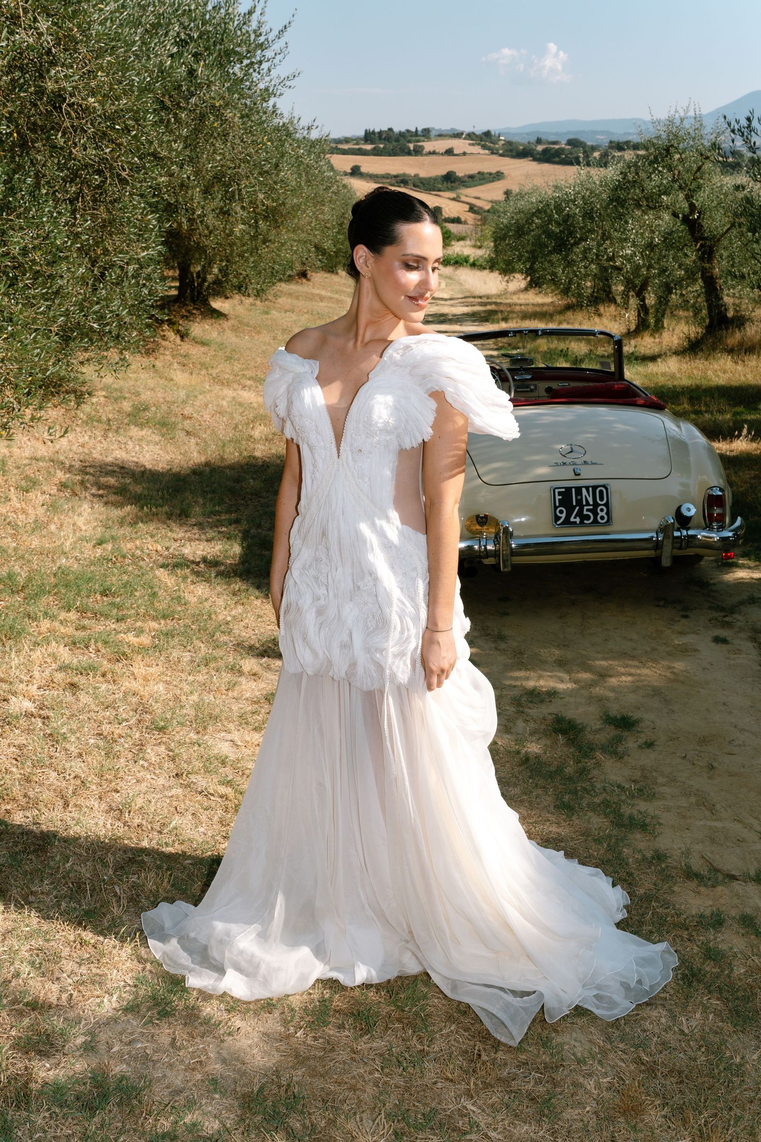 White flowing wedding dress captured in outdoor countryside setting with vintage car in background.