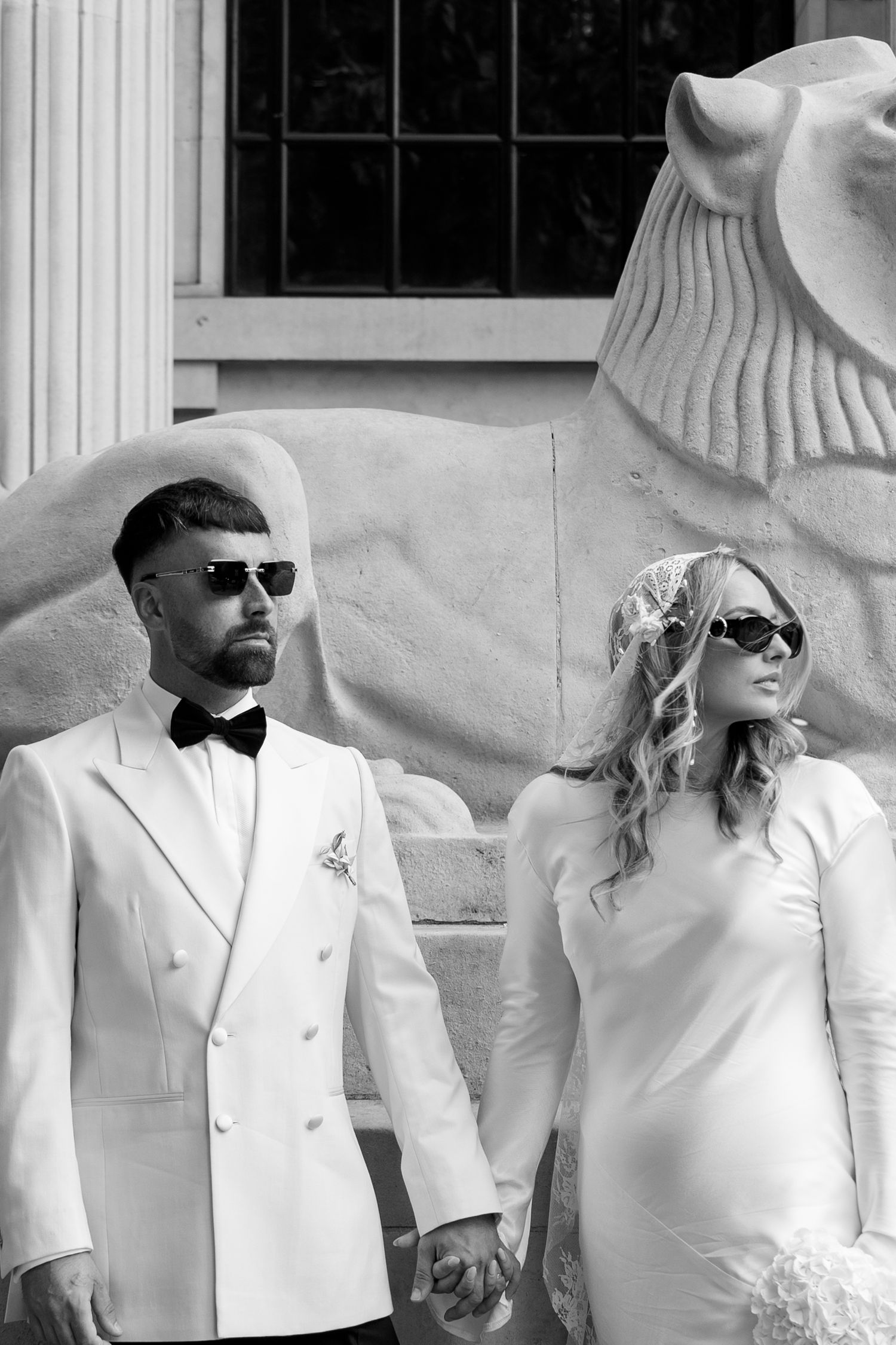 Stylish couple in white attire and sunglasses pose elegantly against a stone backdrop with classical columns.