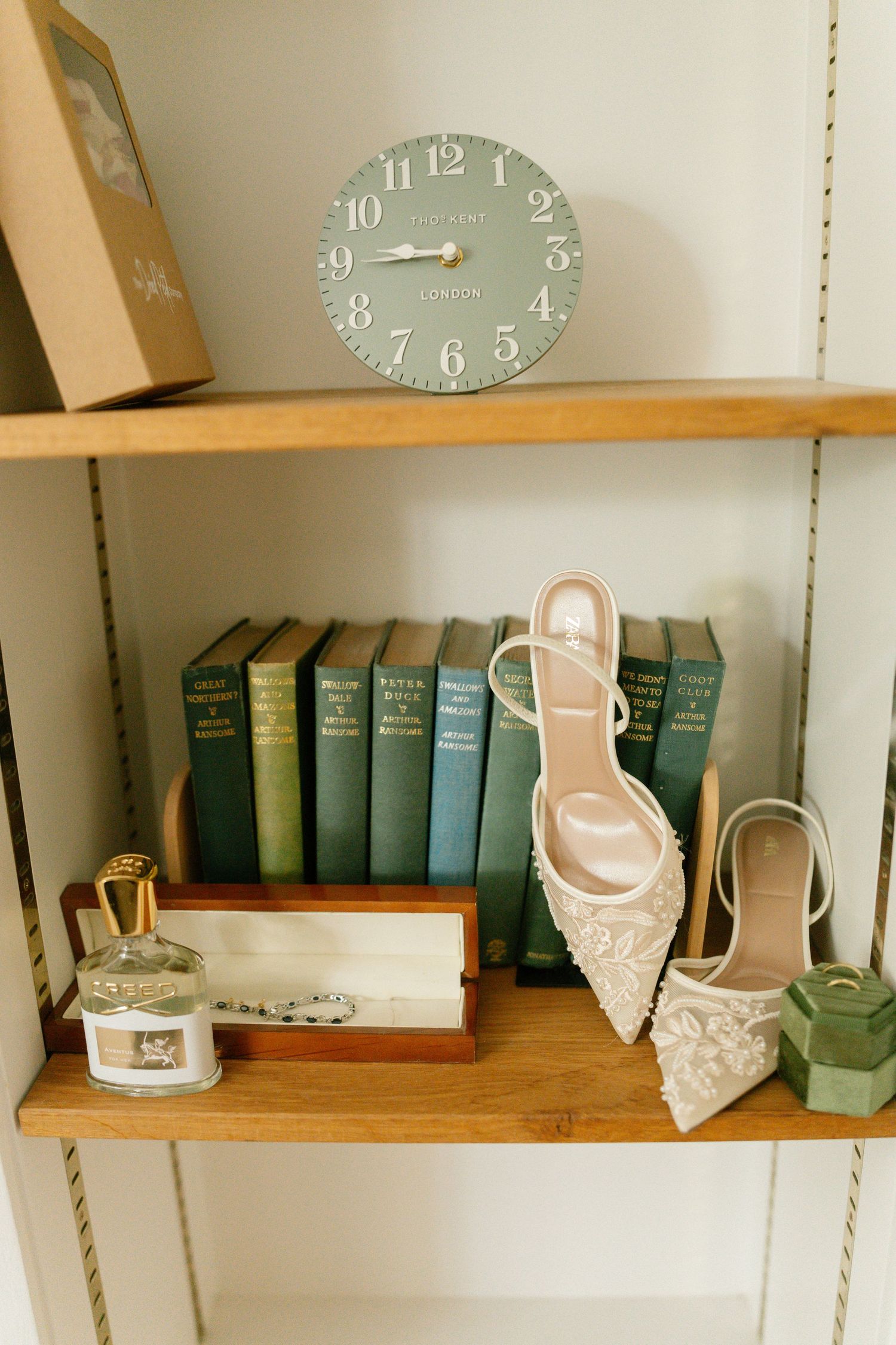 Rustic wooden shelves display vintage books, elegant bridal shoes, and a sage green wall clock in a wedding preparation space.