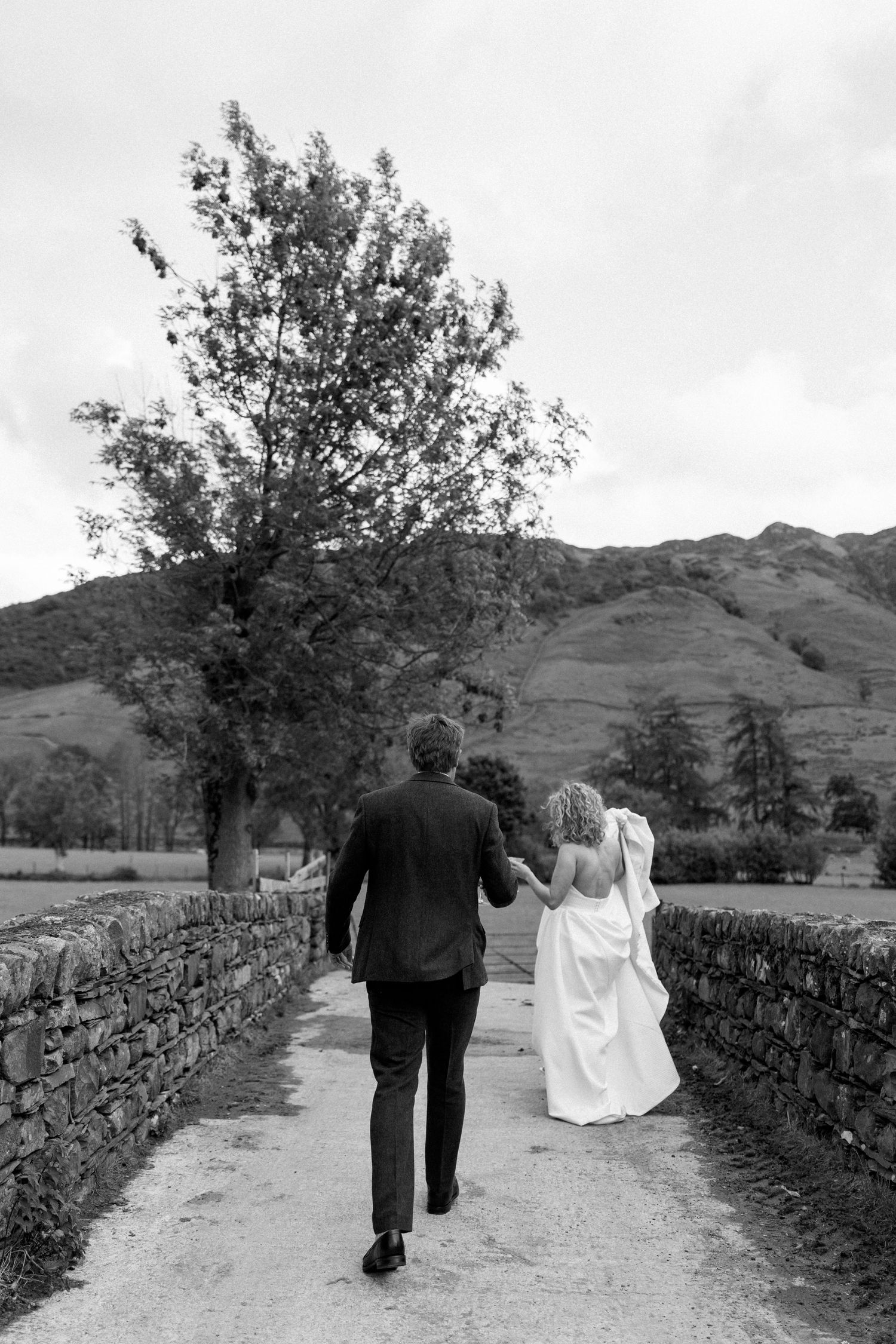 A couple walks along a scenic country path bordered by stone walls with mountains in the background.