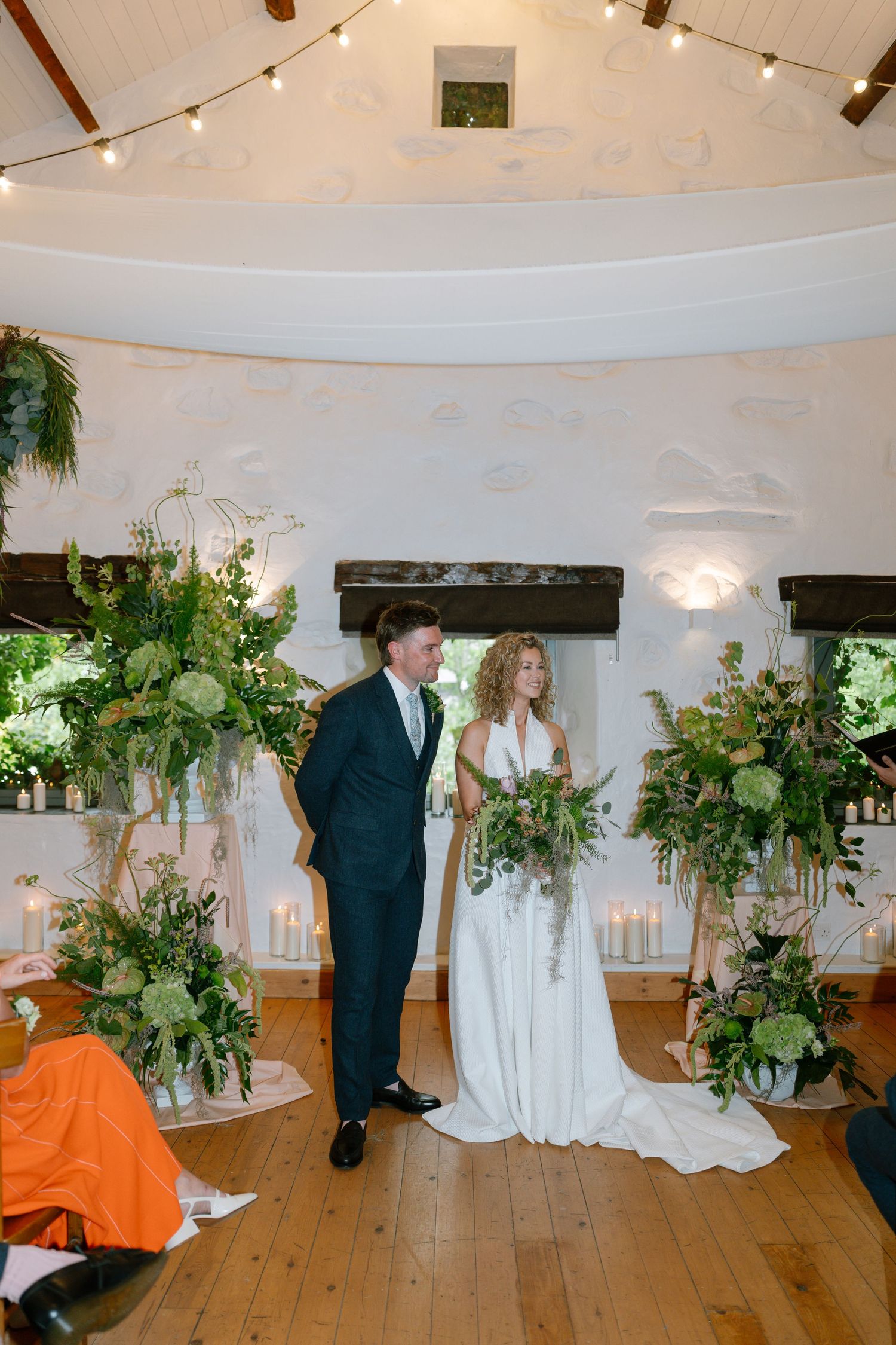 Wedding ceremony setup in a rustic white room with string lights and lush green floral arrangements.