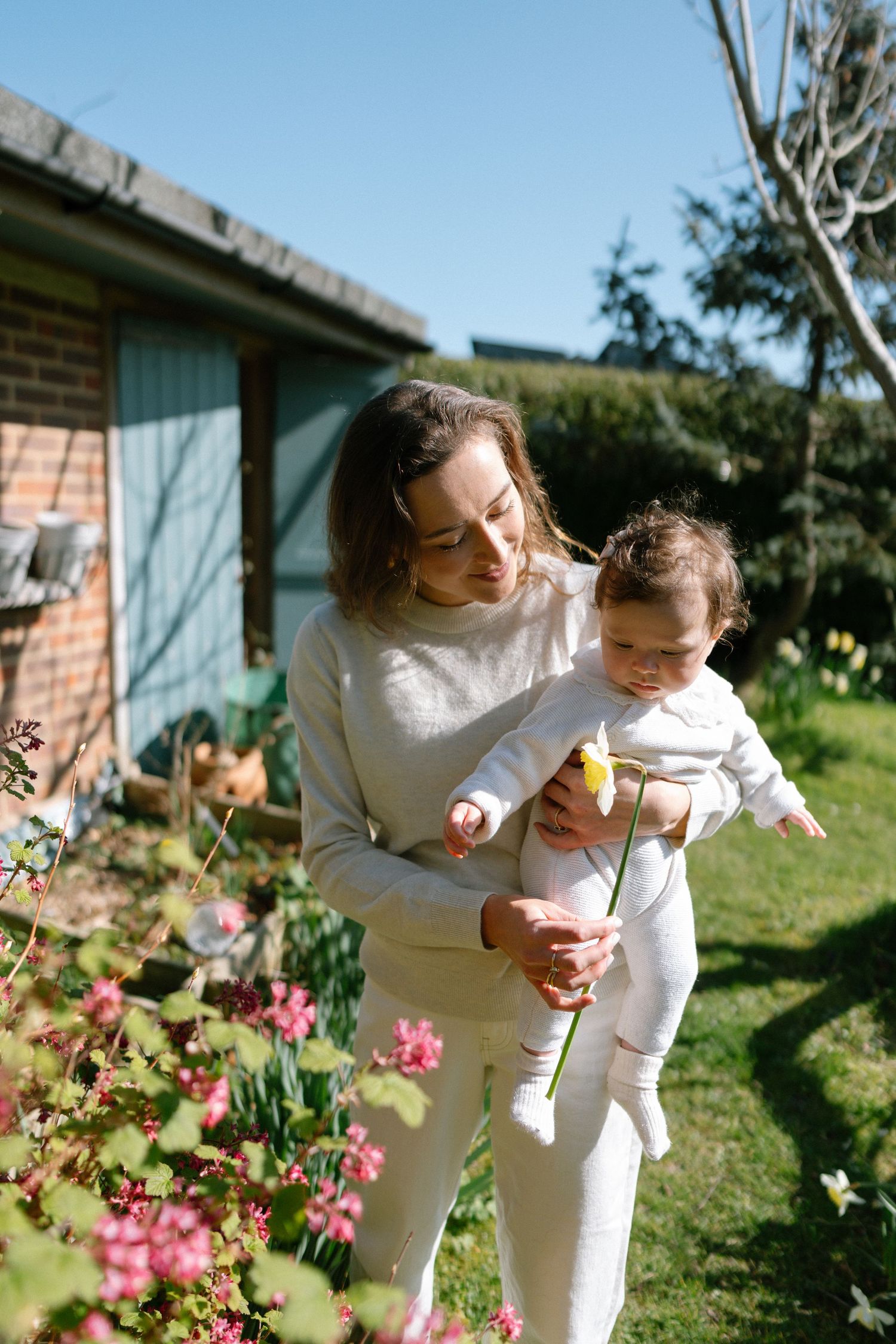 Mother and baby in matching white outfits playing in garden with pink flowers.