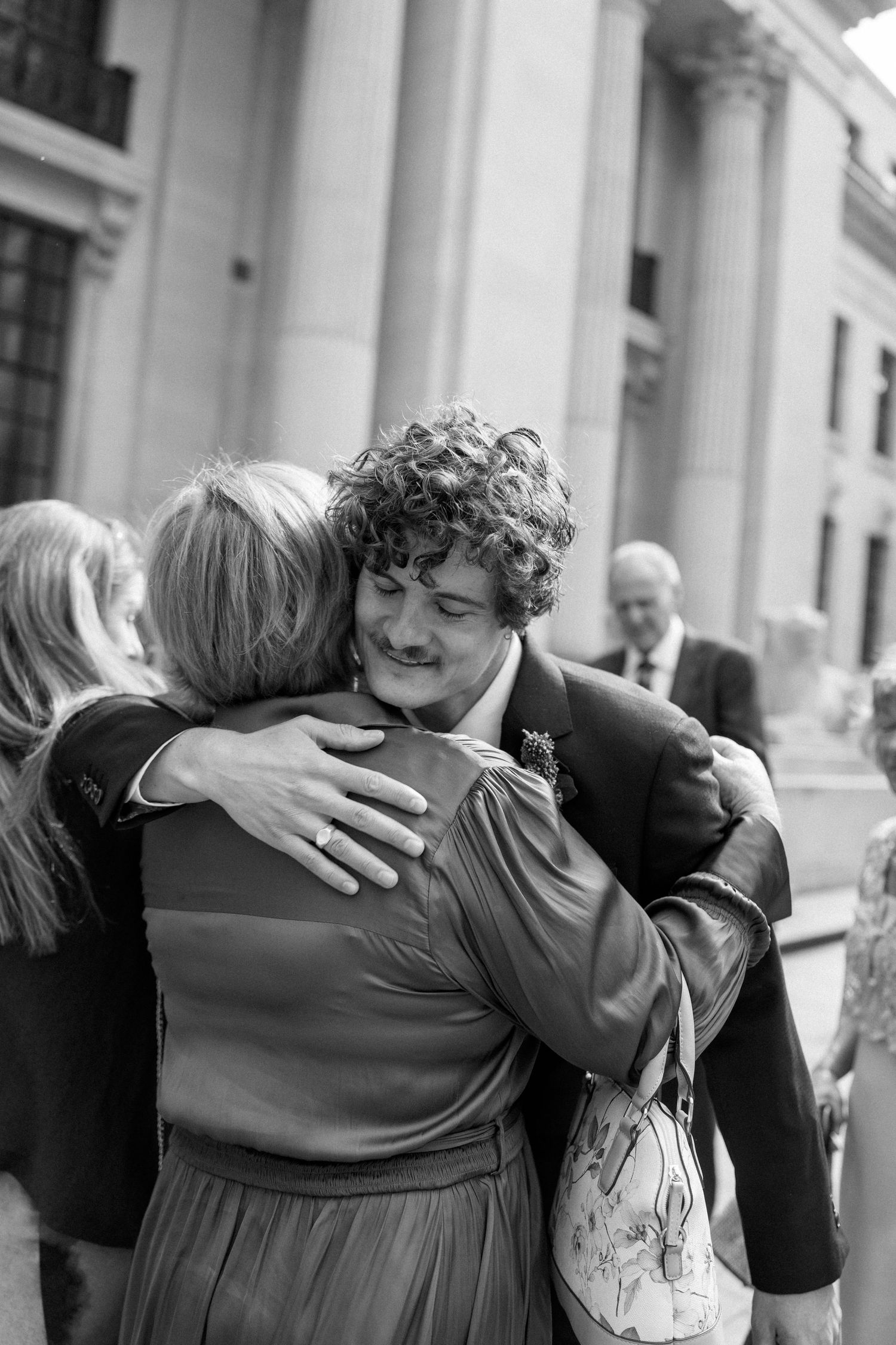 Warm embrace shared between wedding guests outside historic venue.