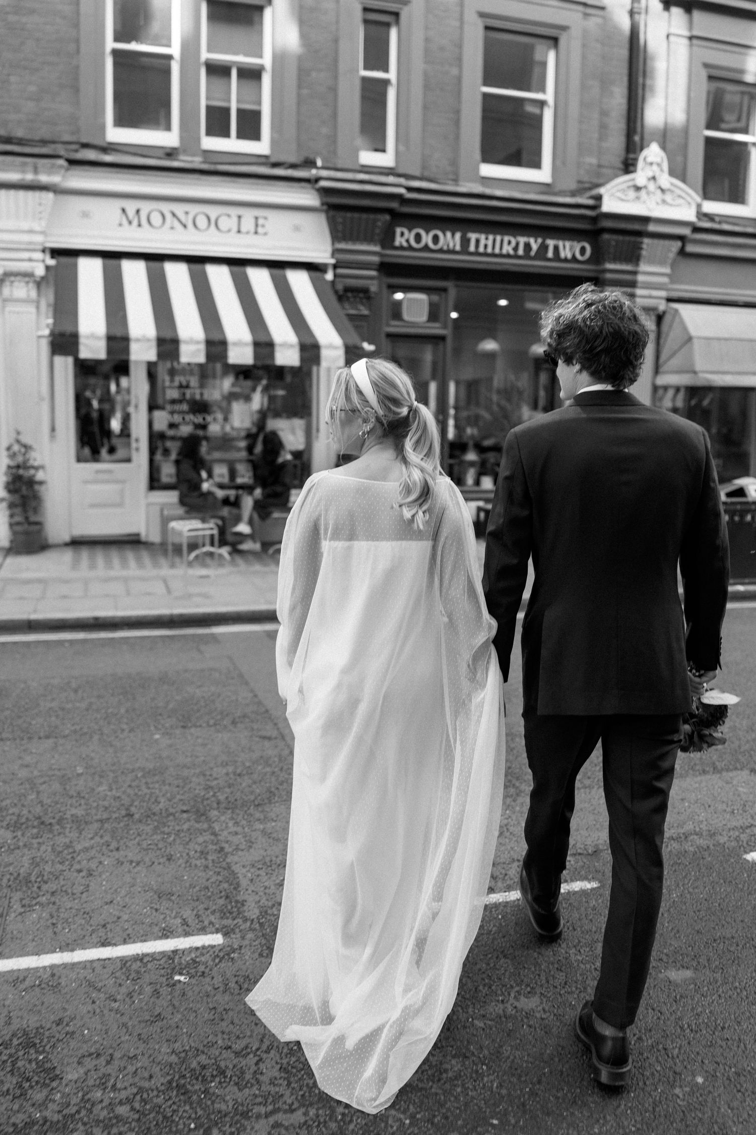 Black and white photo of a wedding couple walking down a city street past retail shops.