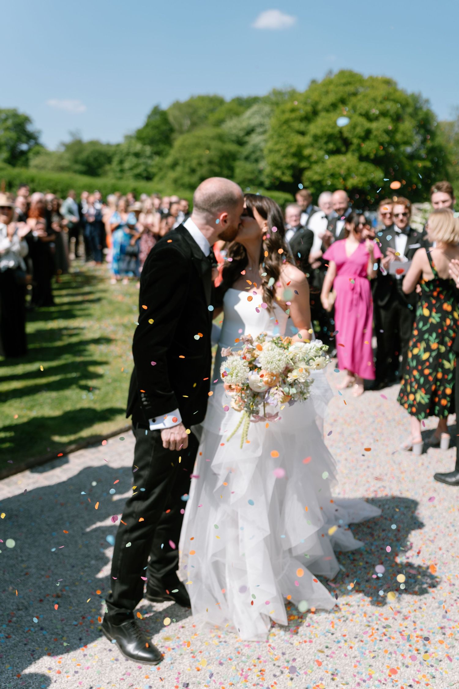 Wedding party walks along a garden path while guests shower them with colorful confetti.