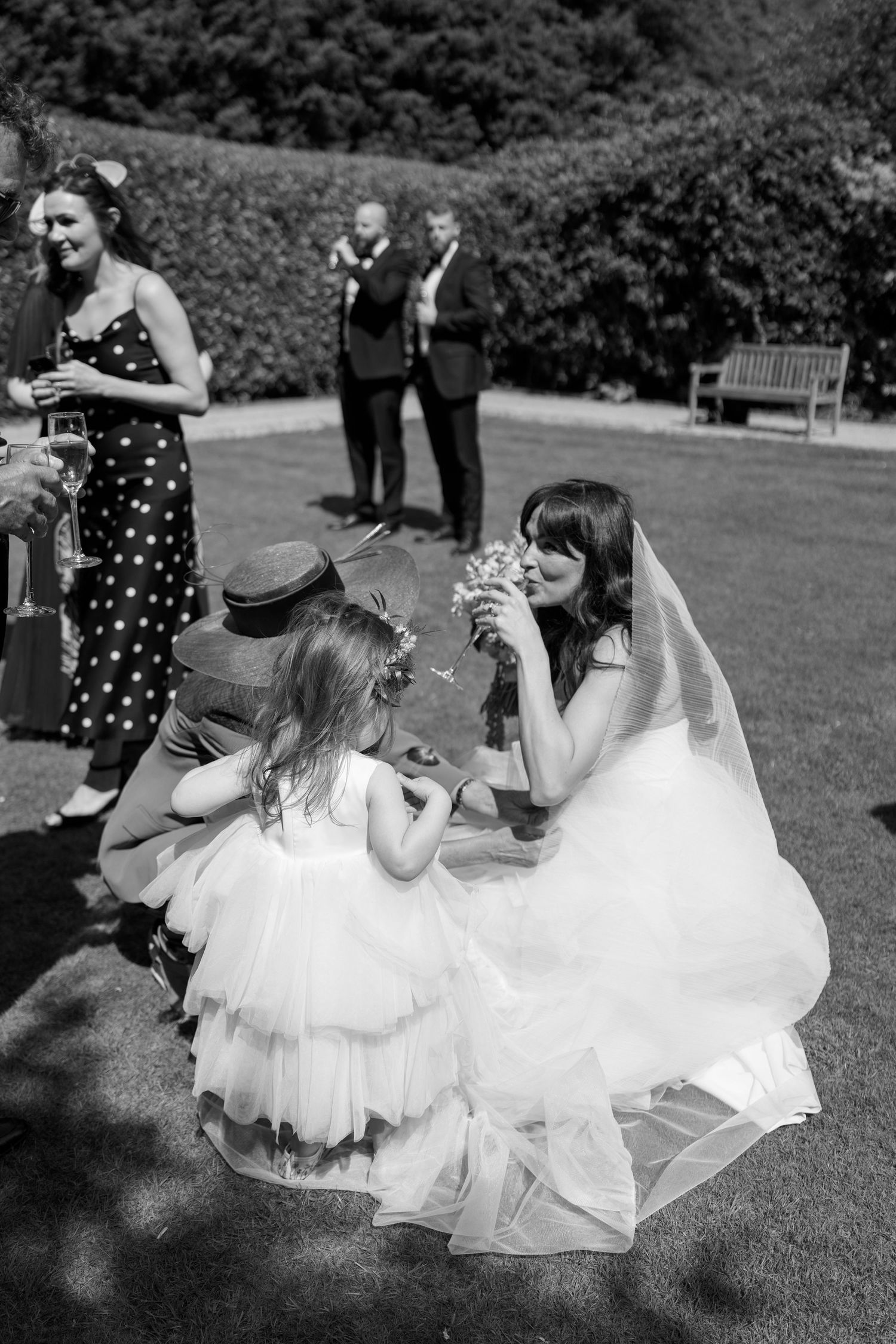 A bride in white dress shares a sweet moment with a flower girl on the lawn.
