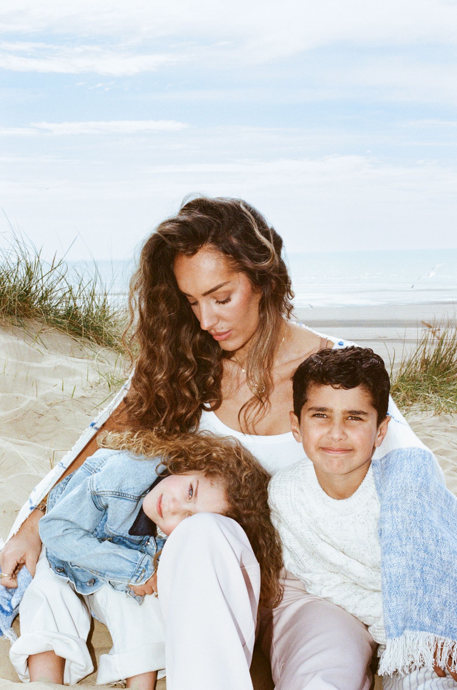 A woman with curly hair sits on a sandy beach with two children, sharing a warm and tender family moment.