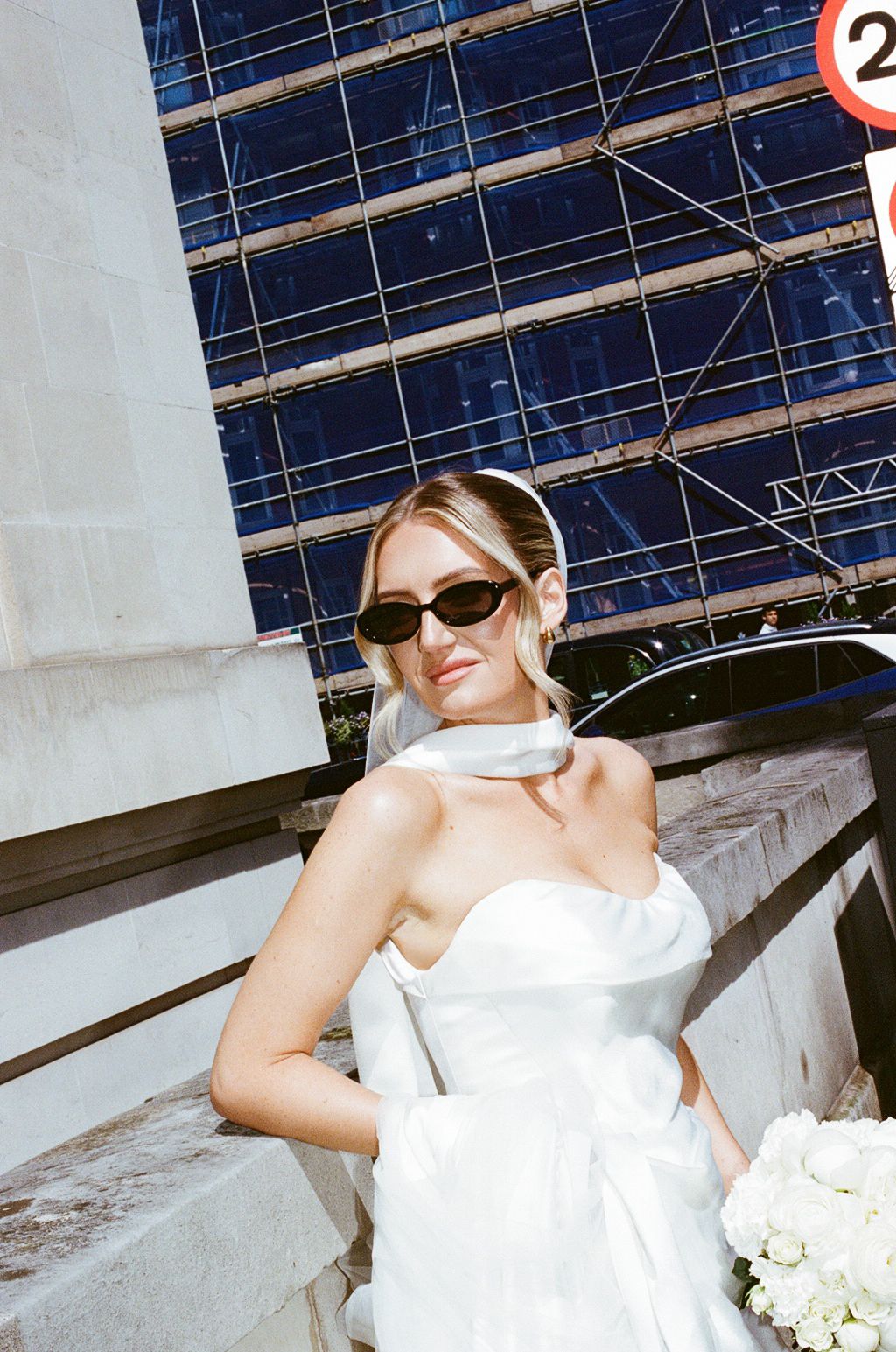 Bride in strapless white gown with veil and sunglasses holding bouquet on urban steps.