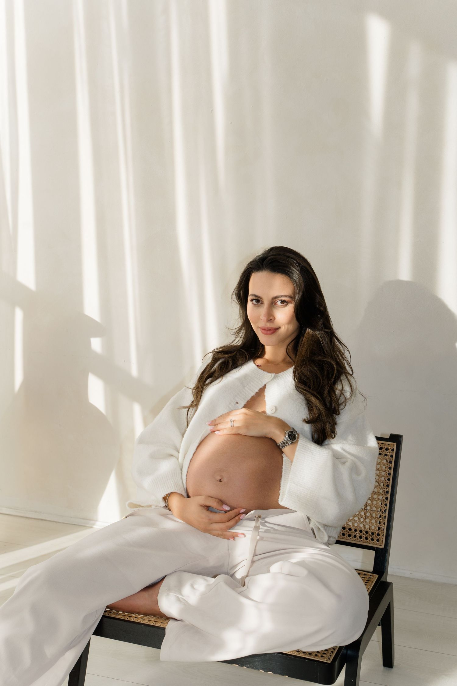 Pregnant woman in white outfit sitting on chair, cradling baby bump in sunlit room with white curtains.