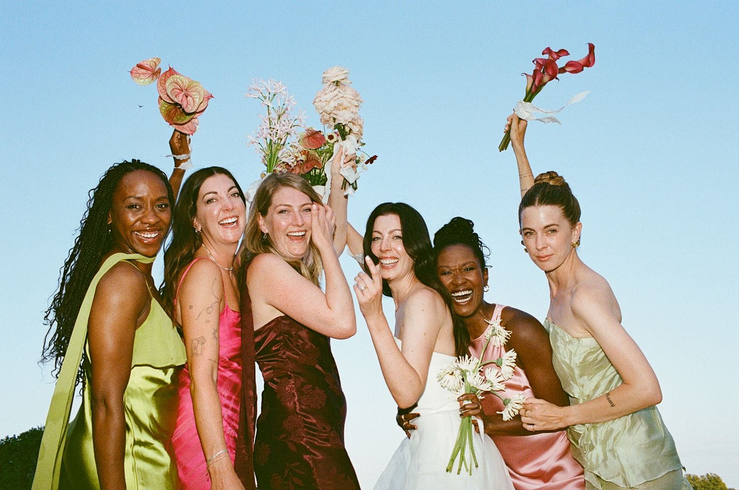 Six bridesmaids in colorful dresses joyfully raise floral bouquets against a clear blue sky at an outdoor wedding.